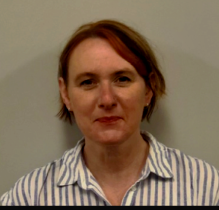 Portrait of a woman with short red hair, wearing a striped collared shirt, standing against a plain wall.
