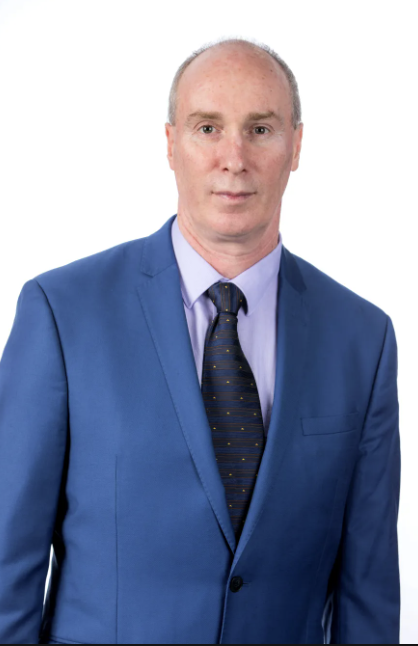 A middle-aged man with a bald head, wearing a blue suit, light blue shirt, and navy tie with small yellow dots, standing against a plain white background.