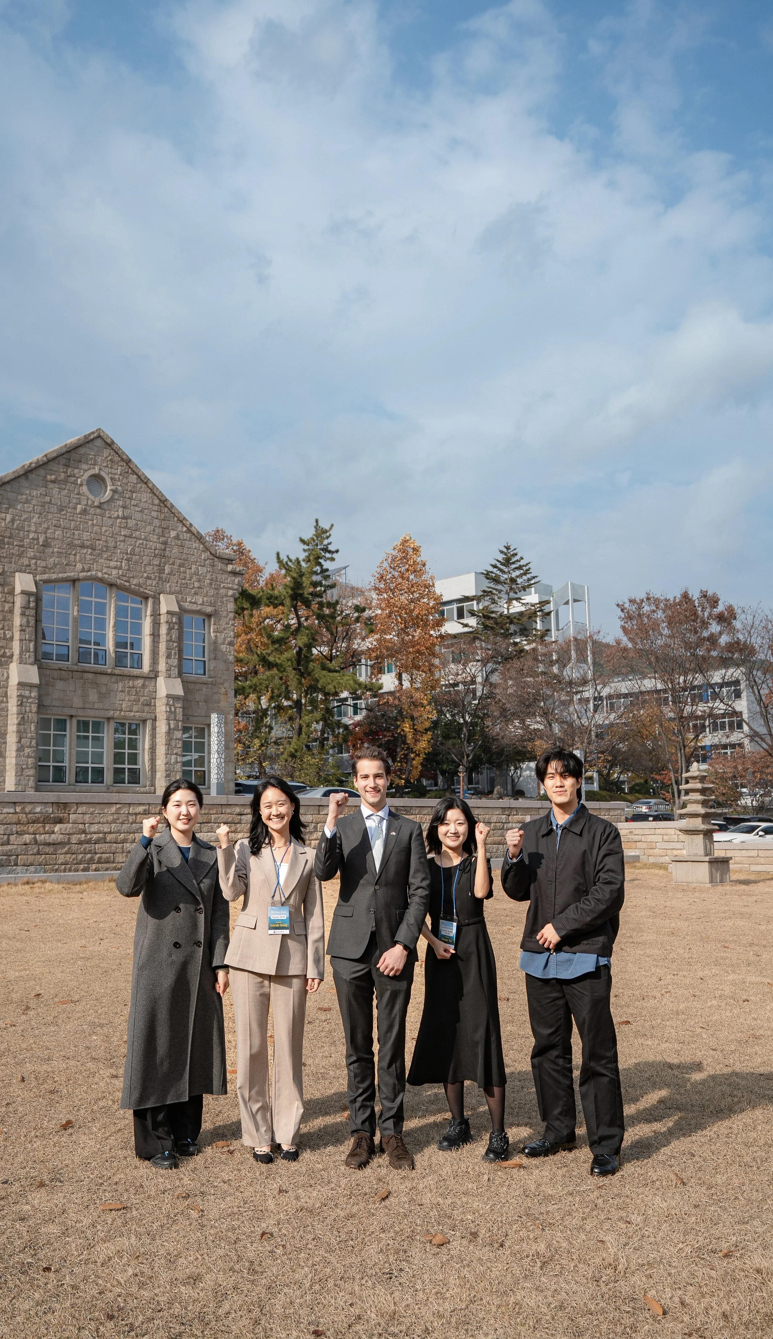 Group of five diverse young professionals standing outdoors on a grassy area, smiling and raising their fists in a gesture of success, with trees and buildings in the background.
