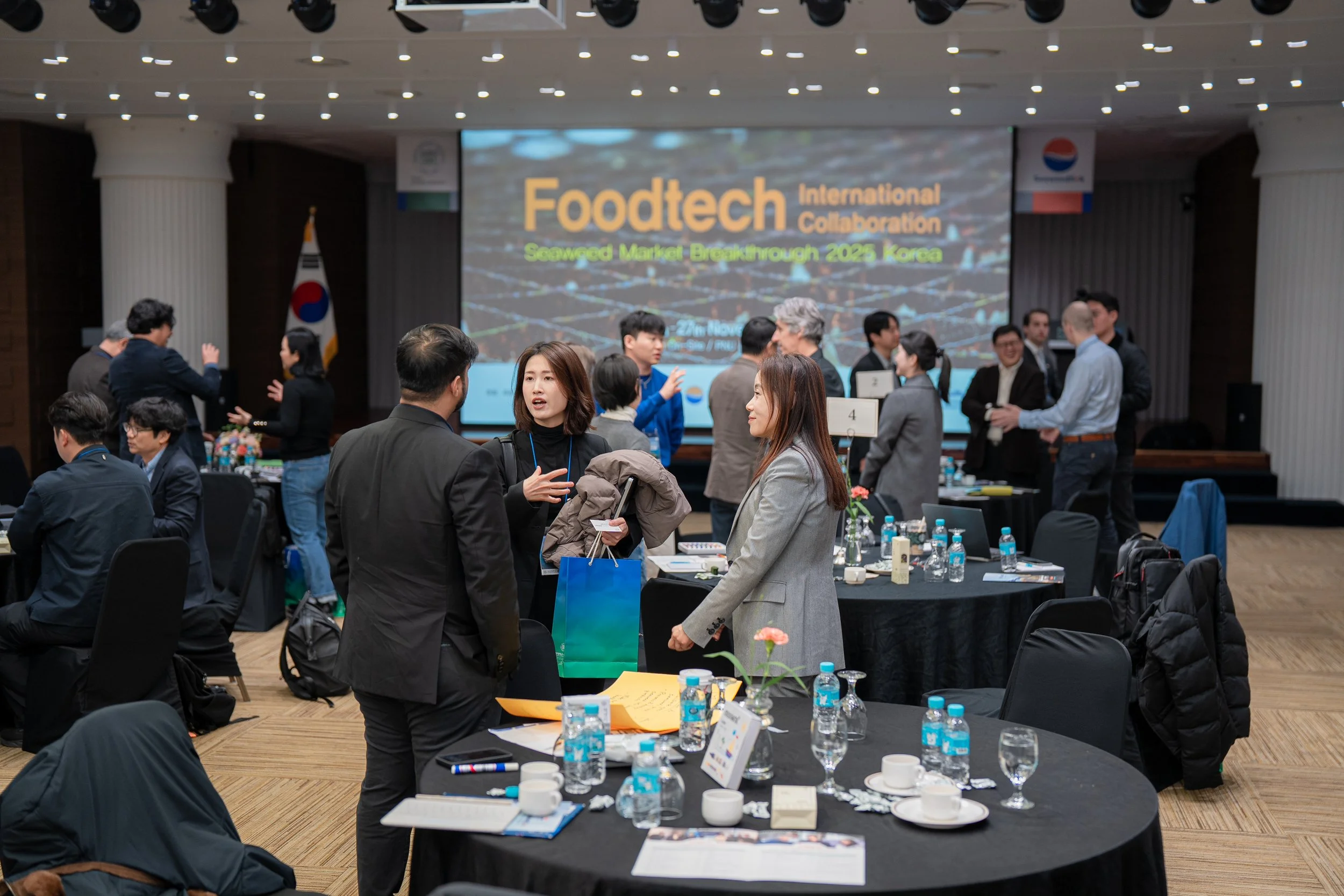 Business conference room with multiple groups of people engaging in discussions, tables with water bottles, and a large screen displaying 'Foodtech International Collaboration' in the background.