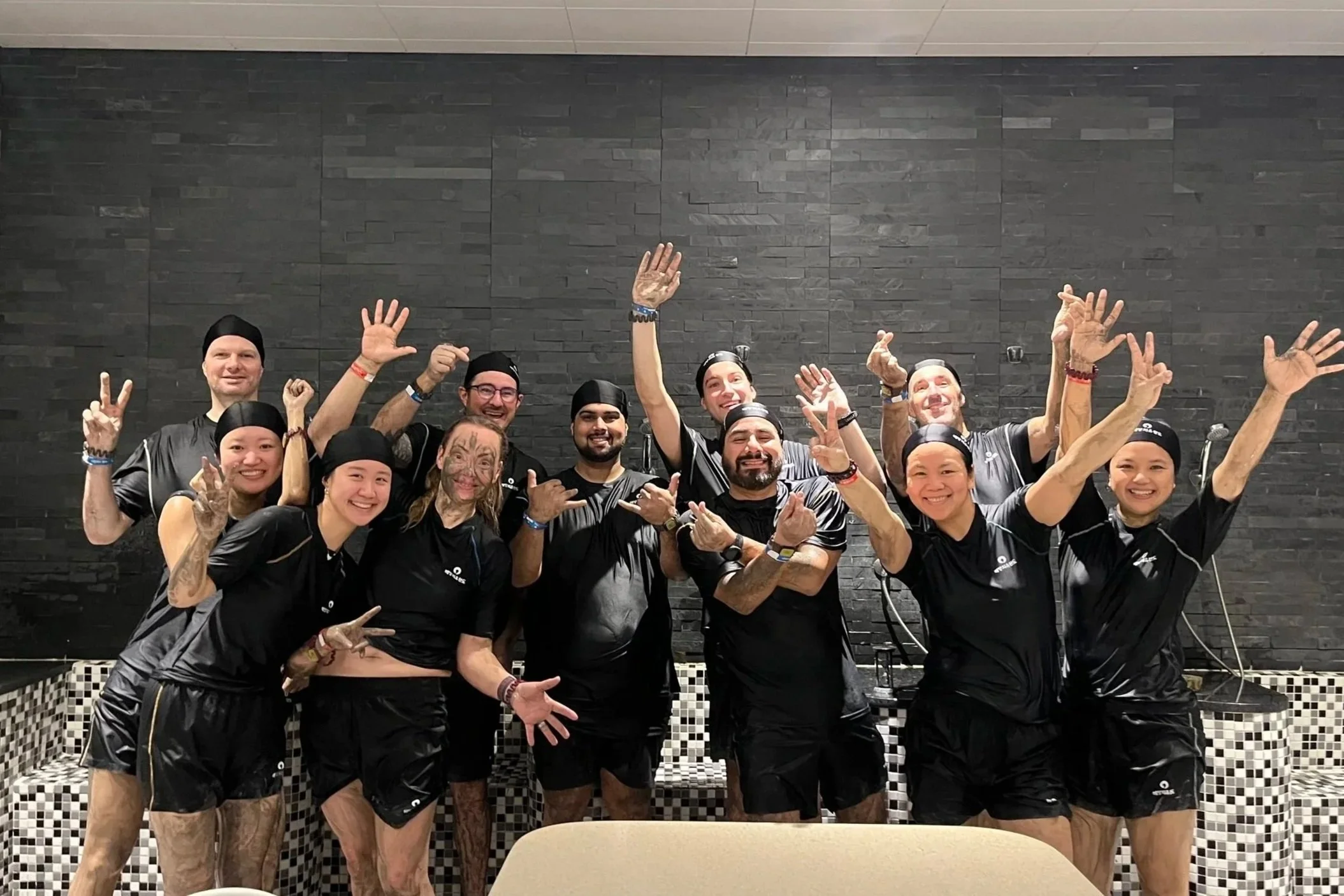 Group of eleven diverse individuals wearing black uniforms, celebrating and posing together in front of a black brick wall.