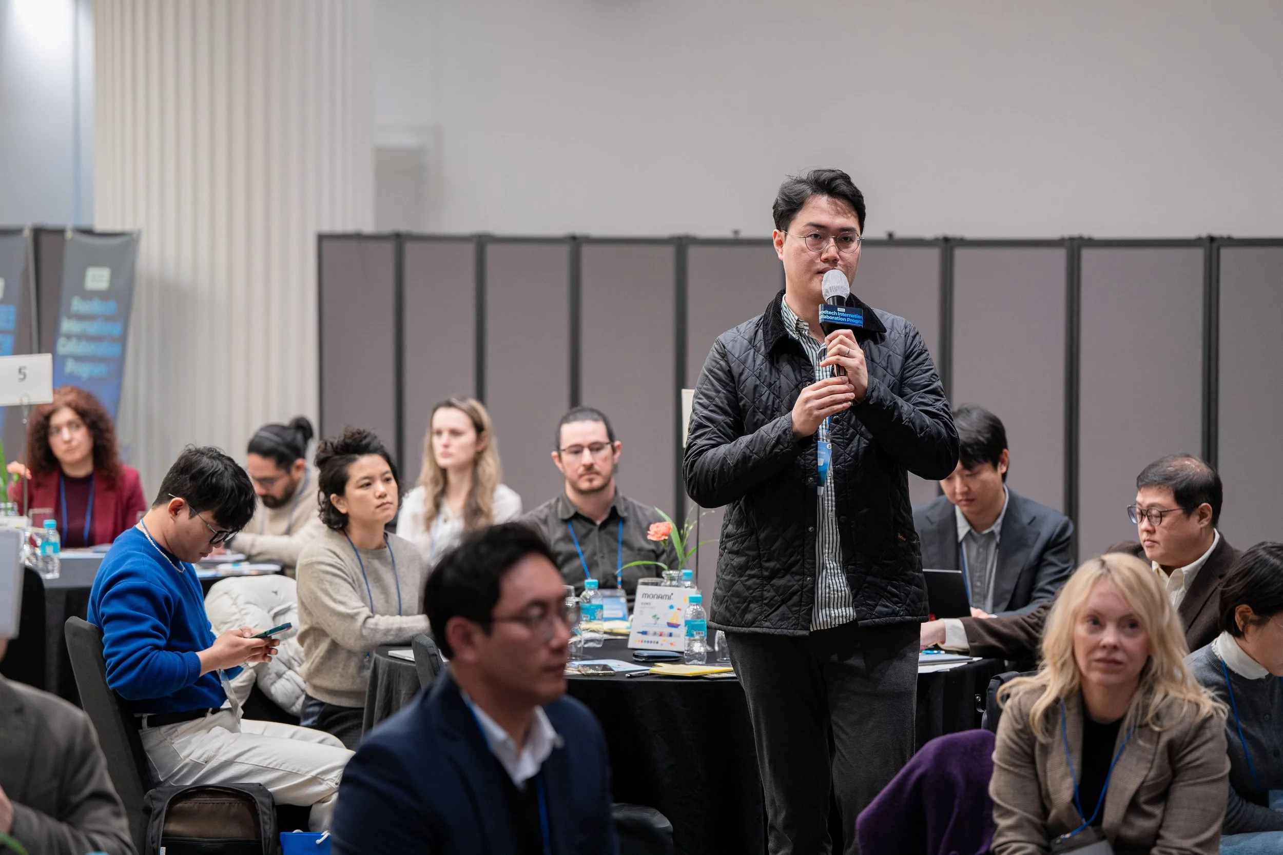 A man standing and speaking into a microphone during a conference, surrounded by seated attendees taking notes or looking at their devices in a large room with informational banners and tables.