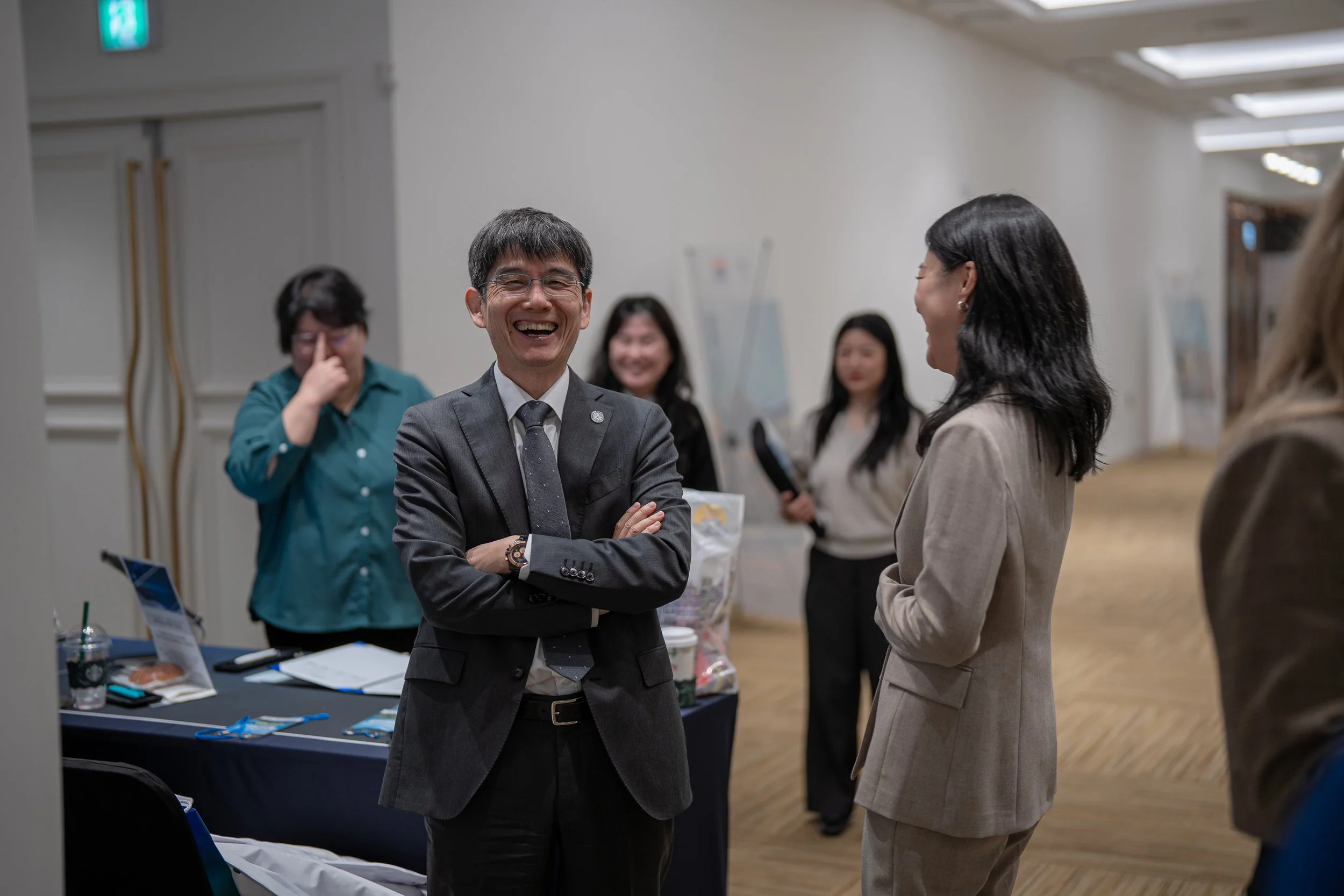 A group of five people in a professional setting, laughing and talking. The man in the foreground is wearing a suit and glasses, standing with crossed arms. Two women are behind him, one in a beige suit and the other holding a microphone. The room has white walls, wooden floors, and a table with event materials.