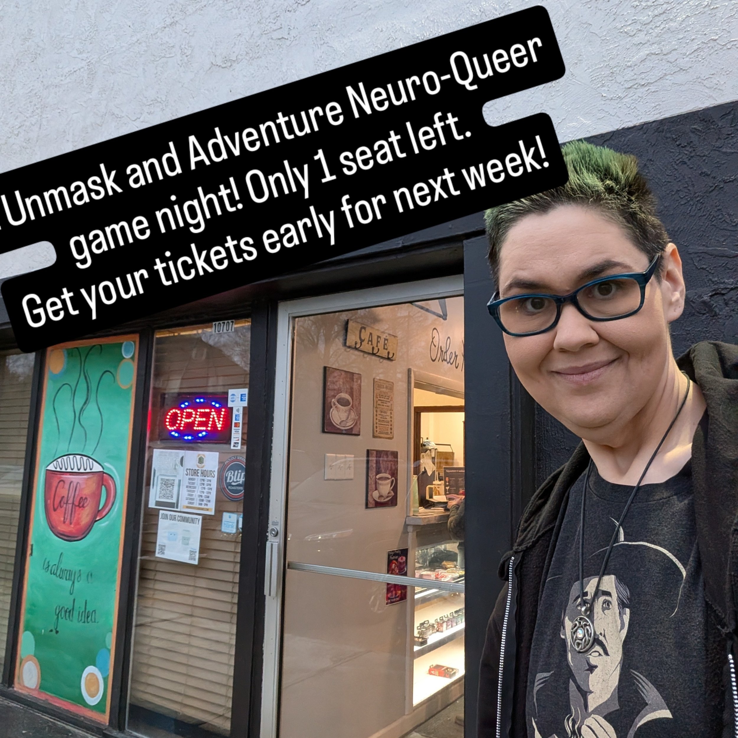 A person taking a selfie outside a cafe, with a sign indicating one one seat left for D&D game night, and a painted coffee cup on the window. The person is wearing glasses, a black shirt with a graphic, and is smiling.