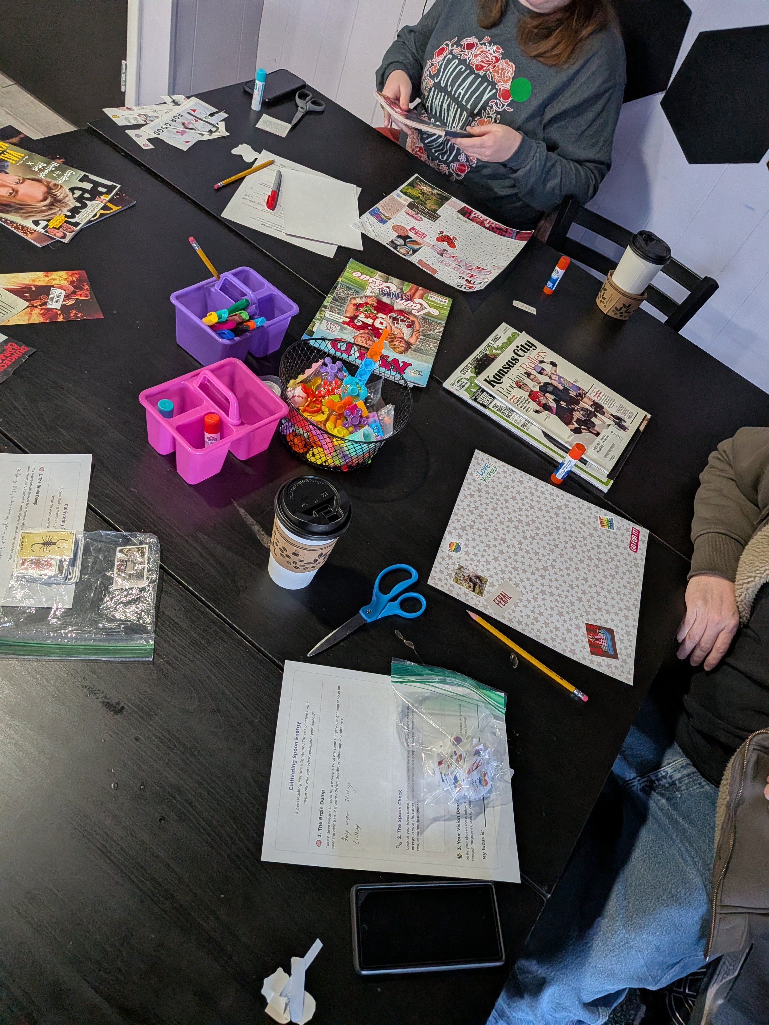 A black table with various crafting supplies, magazines, and papers. There are small containers with markers, cutouts, glue sticks, scissors, a cup of coffee, and a person holding a magazine. The table is in a room with white walls.