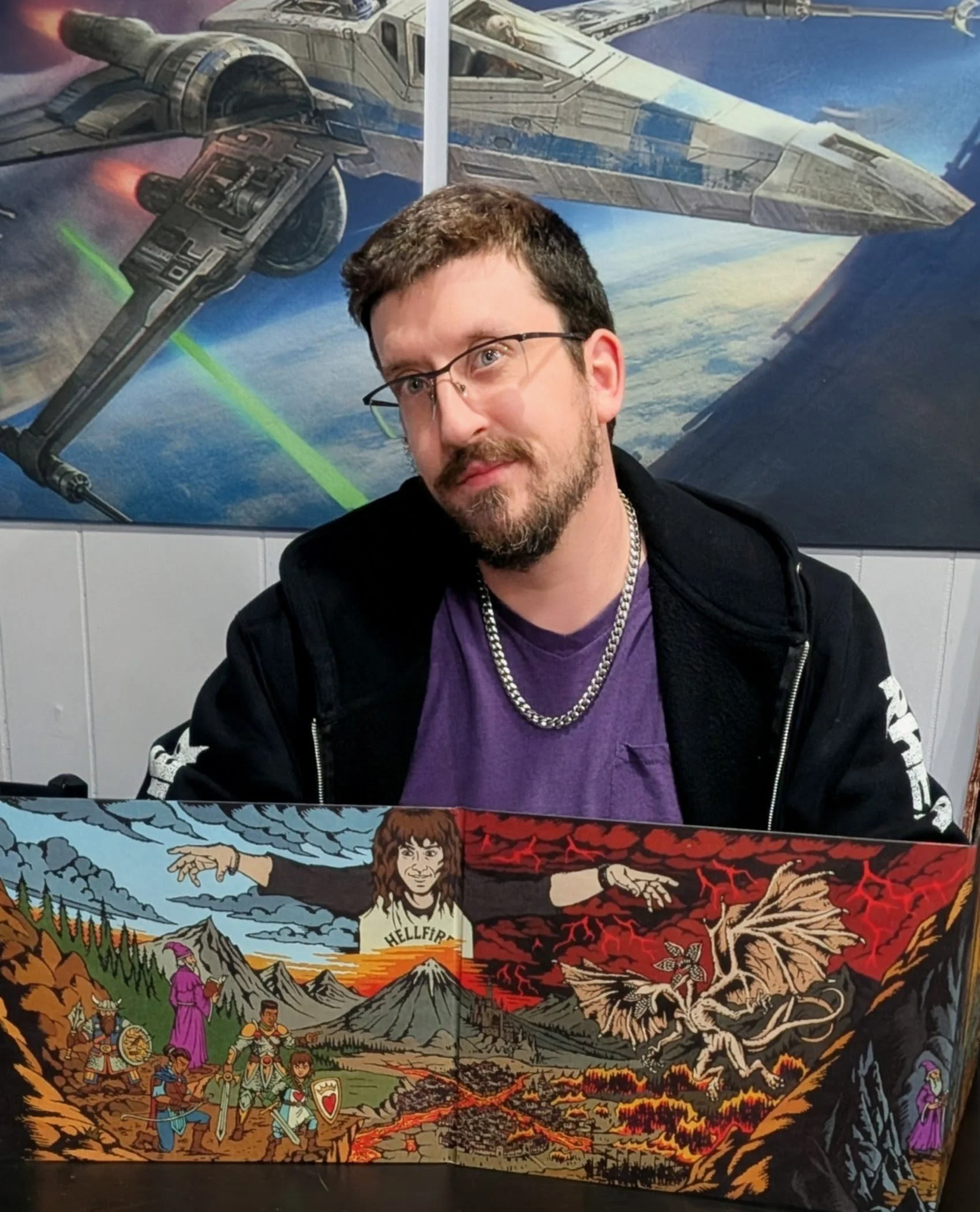 A man with glasses and a beard sits in front of a space-themed poster featuring a spaceship. He is wearing a purple shirt, a black hoodie, and a silver chain necklace, and is holding a colorful comic-style art piece depicting fantasy characters, dragons, and mountains.
