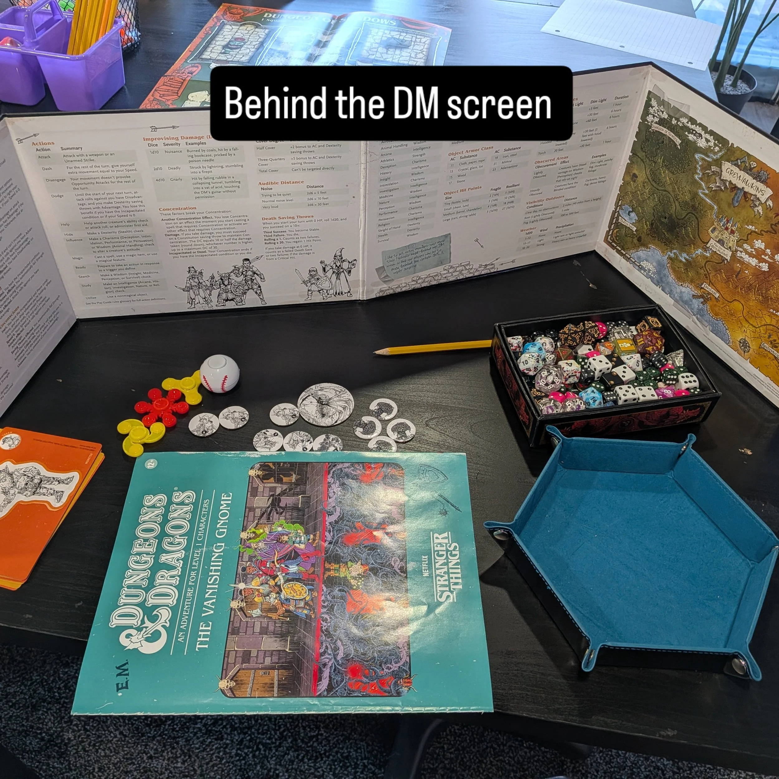 Table with Dungeons & Dragons game materials including rulebooks, tokens, dice, and a dungeon map. A box of colorful polyhedral dice is open on the right, with a blue storage tray nearby. Behind the table, an annotated map and game statistics are visible.