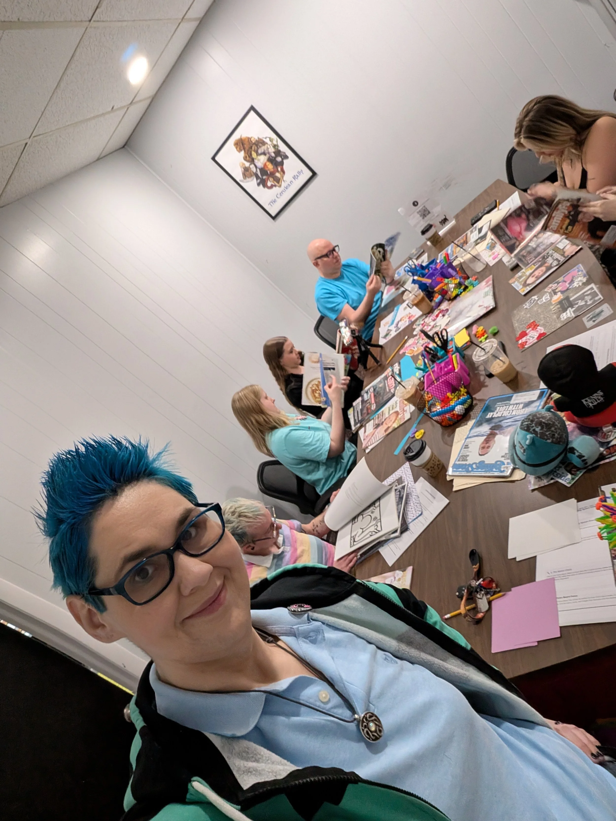 A person with blue hair and glasses taking a selfie at a table with various comics, drawings, and art supplies, surrounded by others reading and working on their projects in a room with white walls and a picture on the wall.