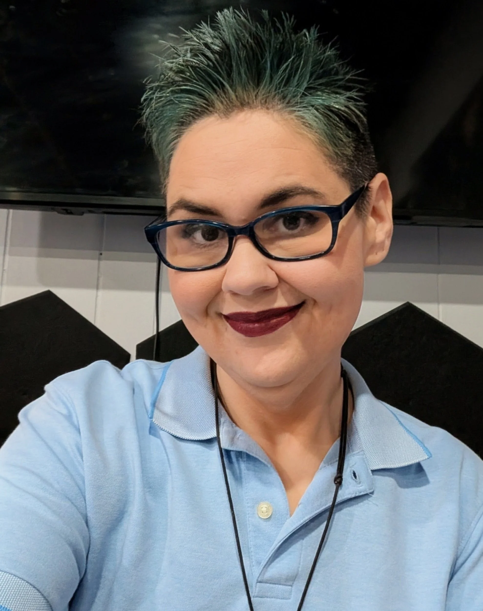 A woman with short hair highlighted in teal, wearing glasses, lipstick, a light blue polo shirt, and a lanyard, smiling at the camera in an indoor setting with black and white geometric decor behind her.