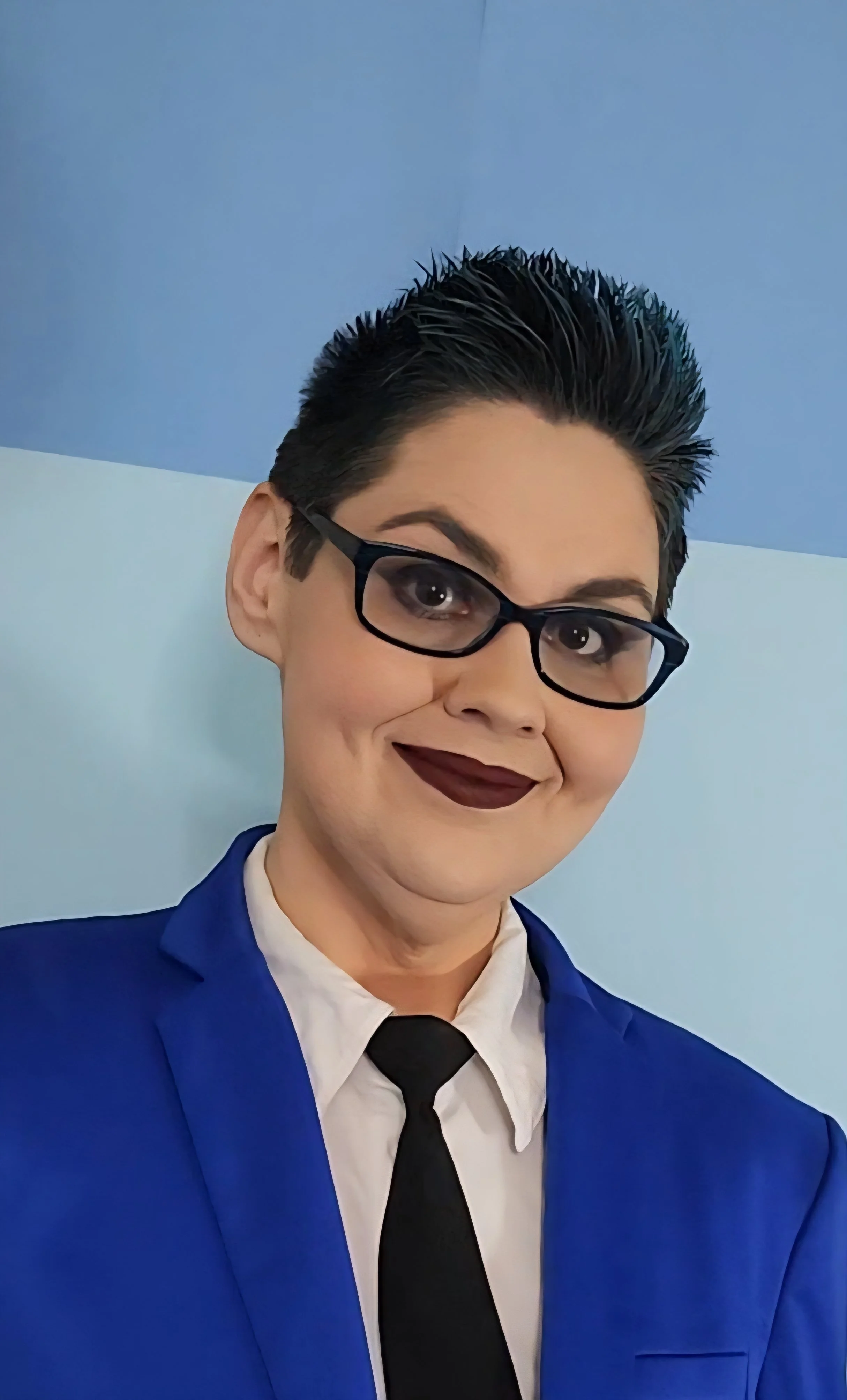 A woman with short, spiked dark hair, glasses, dark lipstick, wearing a blue suit jacket, white shirt, and black tie, smiling against a two-tone blue background.