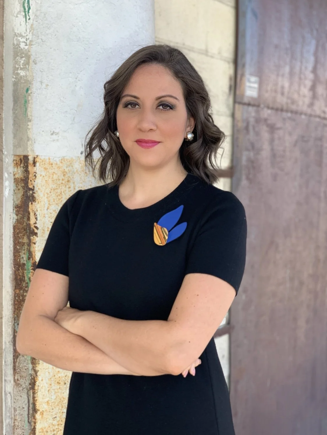 A woman with shoulder-length brown hair, wearing pearl earrings and a black dress with a blue and orange lapel pin, standing with arms crossed in front of a textured wall.