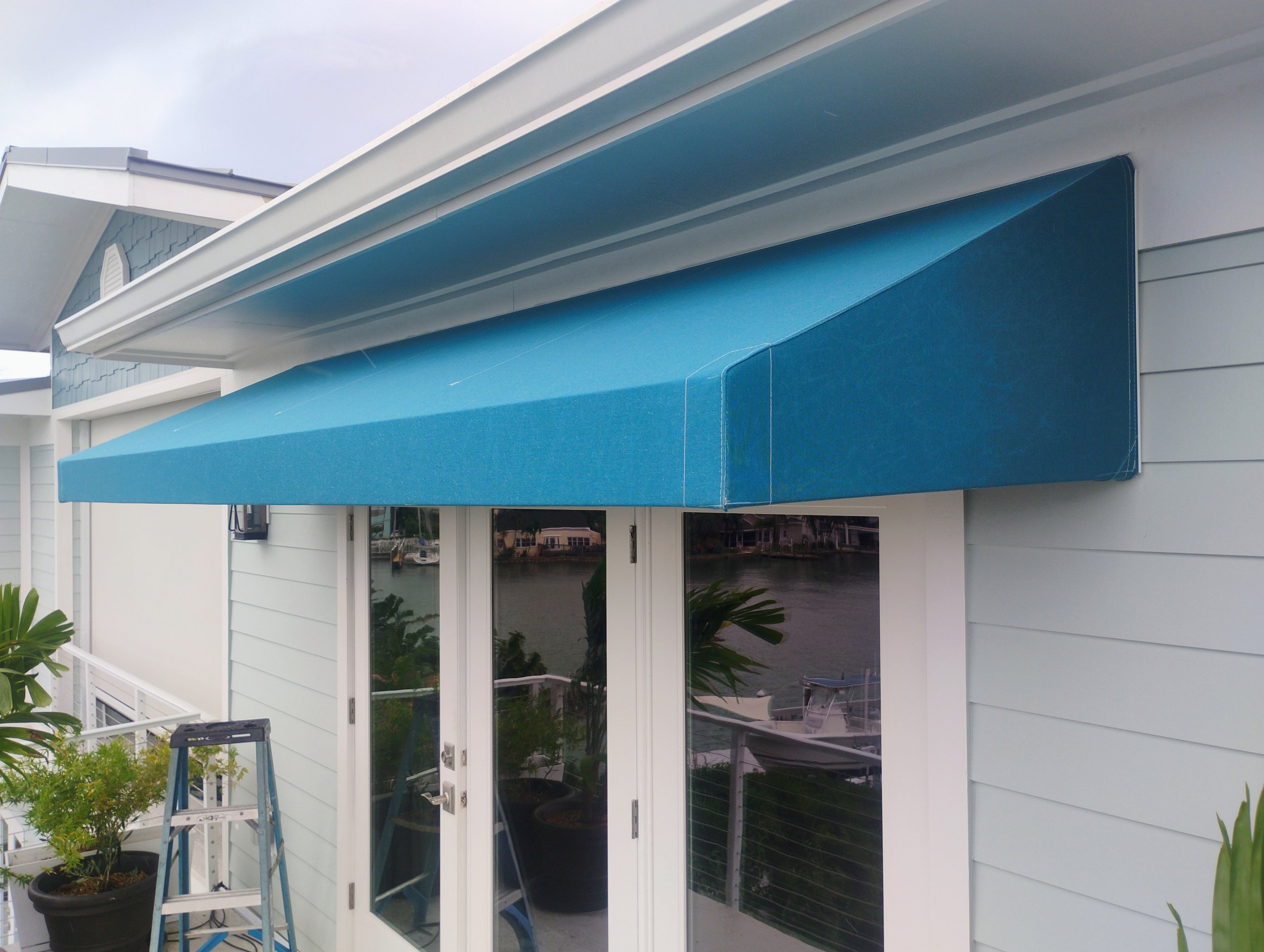 Blue retractable awning over glass doors on a white house near water, with potted plants and a step ladder nearby.