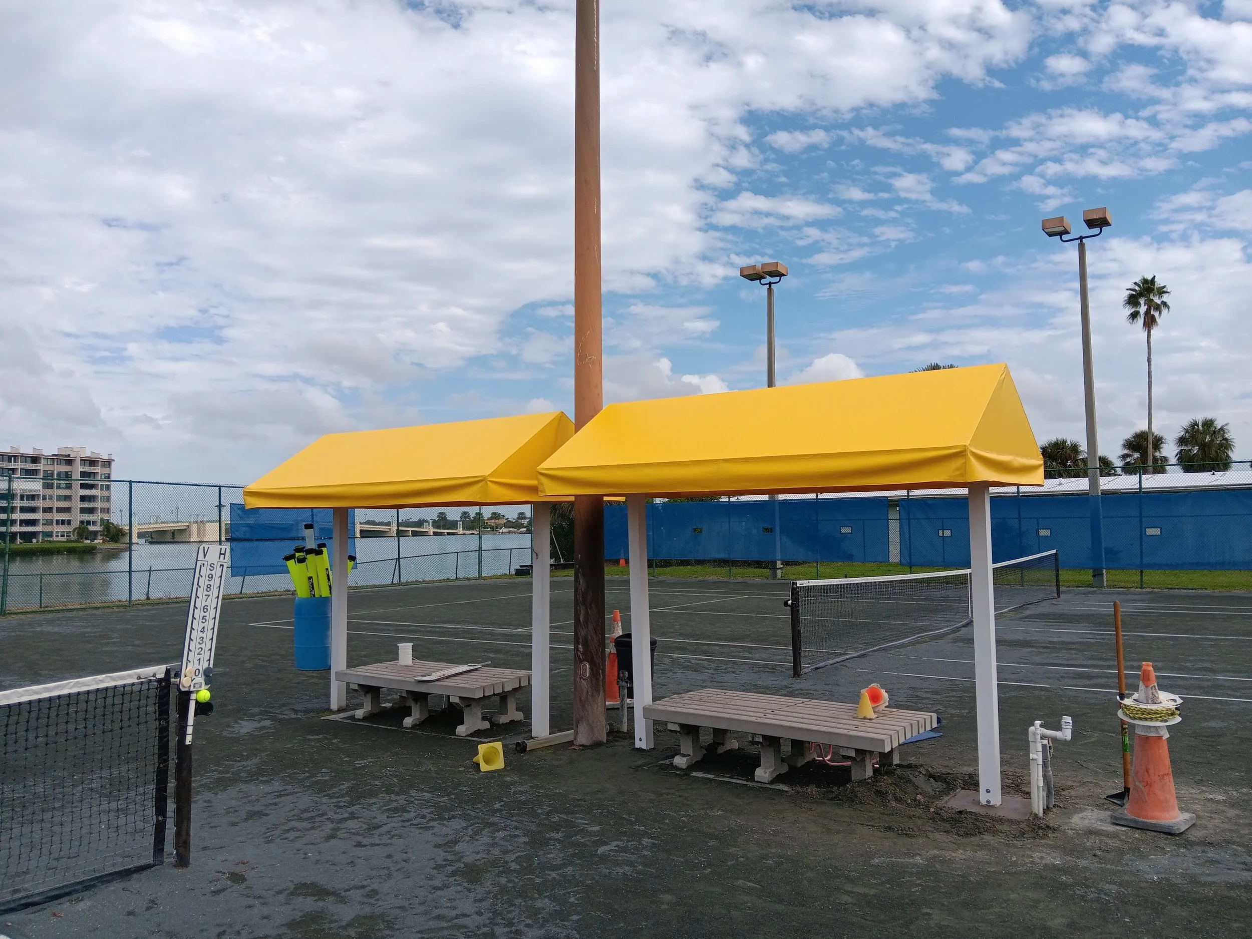 Empty outdoor tennis court with two yellow shade structures, benches, tennis balls, and court equipment under a partly cloudy sky