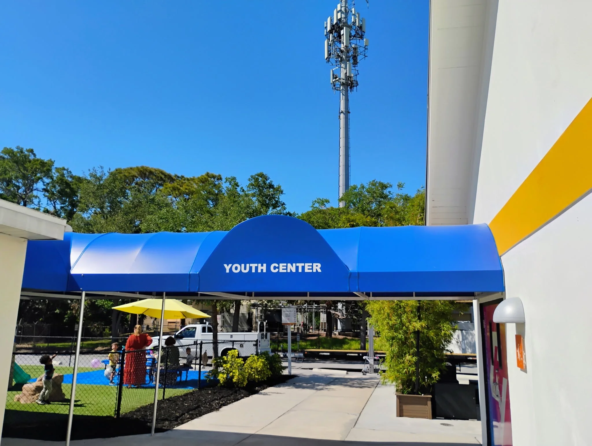 Entrance to a youth center with a blue awning labeled 'YOUTH CENTER.' There are children playing with purple and blue balls on a small playground, supervised by adults, under a yellow umbrella. A tall cell tower is visible in the background, with tre