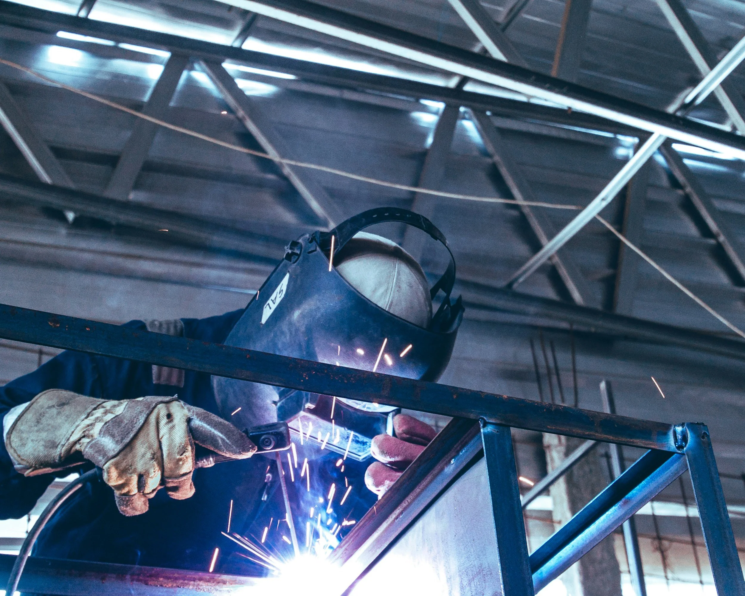 A welder welding metal in an industrial workshop, wearing a protective welding helmet and gloves, with sparks flying from the welding process.