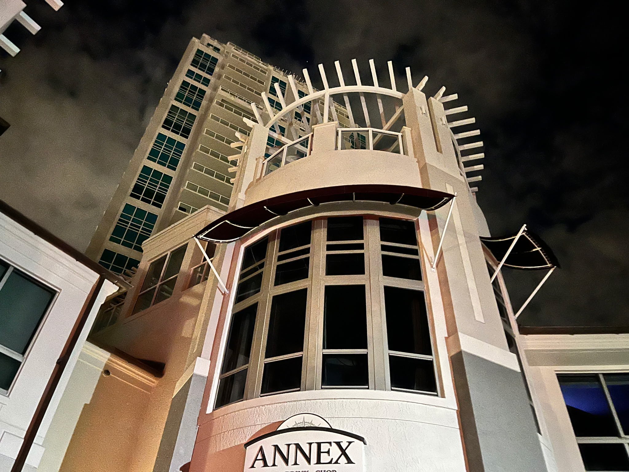 Nighttime view of a modern white building with large windows, decorative architectural features, and a sign reading 'ANNEX' at the bottom, with a taller building in the background and a cloudy dark sky overhead.