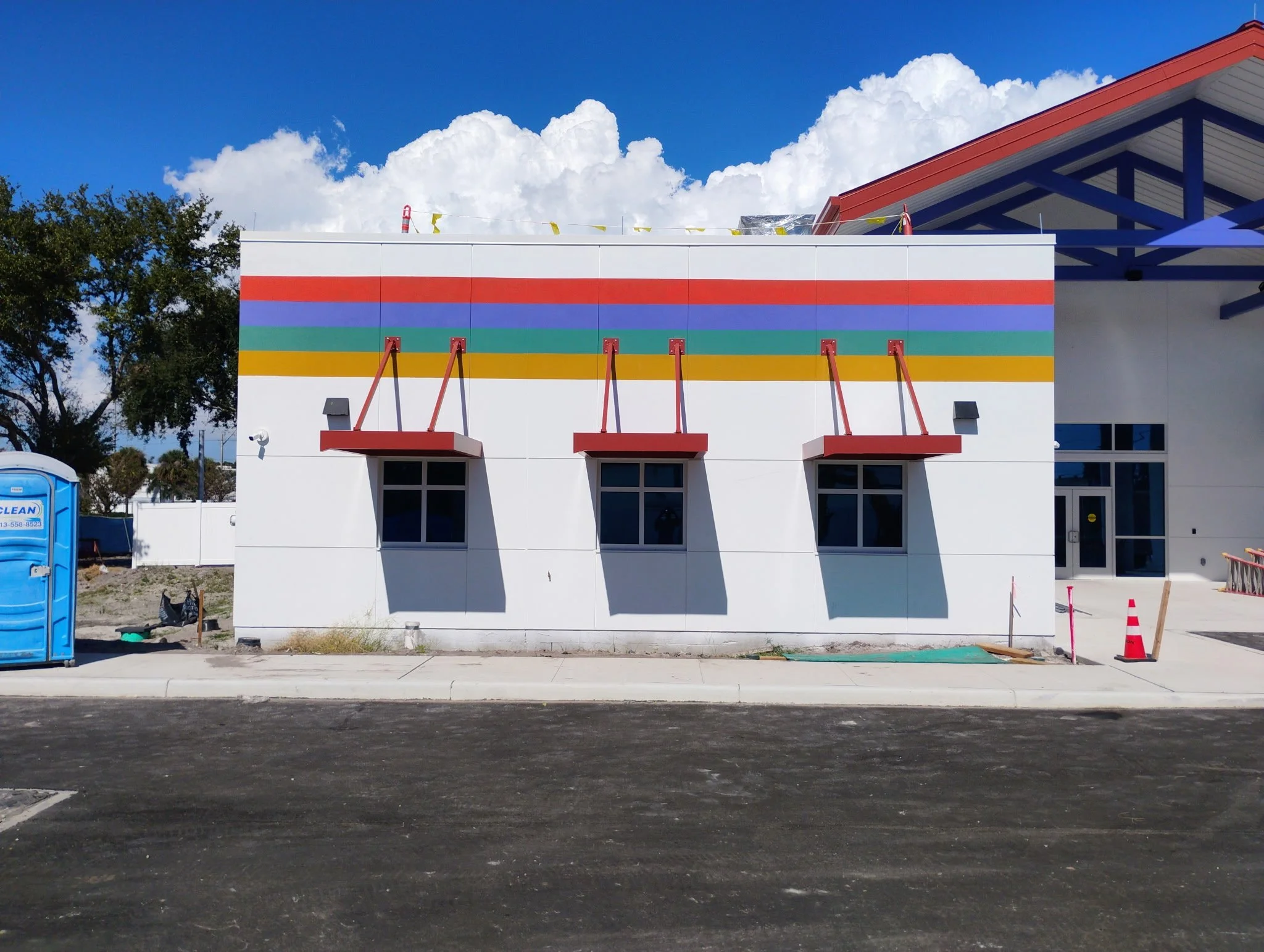A white exterior building with colorful horizontal stripes near the top, three small red awnings over the windows, and a clear blue sky with clouds in the background.