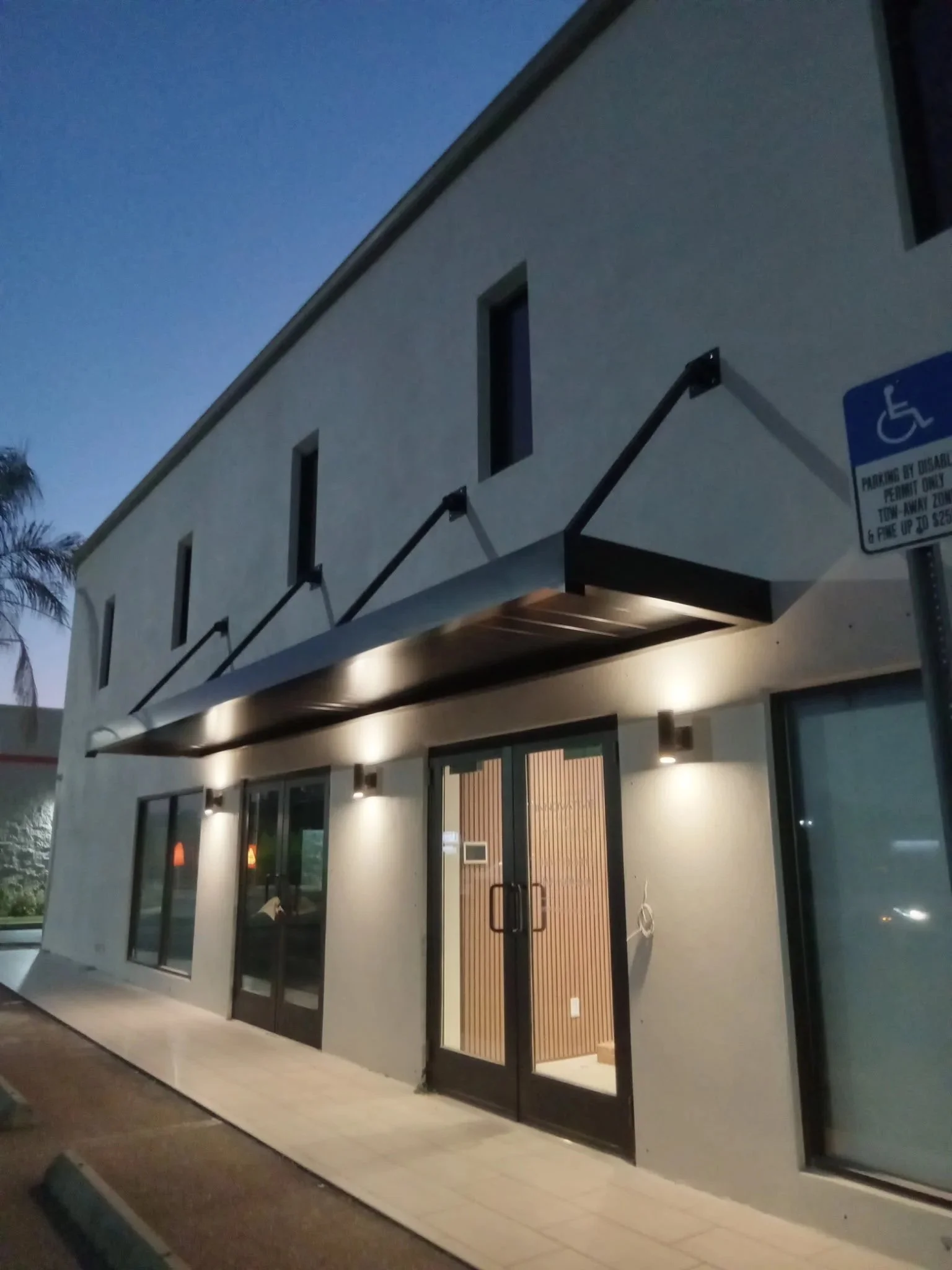 Modern two-story building with a light-colored exterior, glass entrance doors, and a black metal awning illuminated by exterior lights at dusk. Handicap parking sign visible nearby.