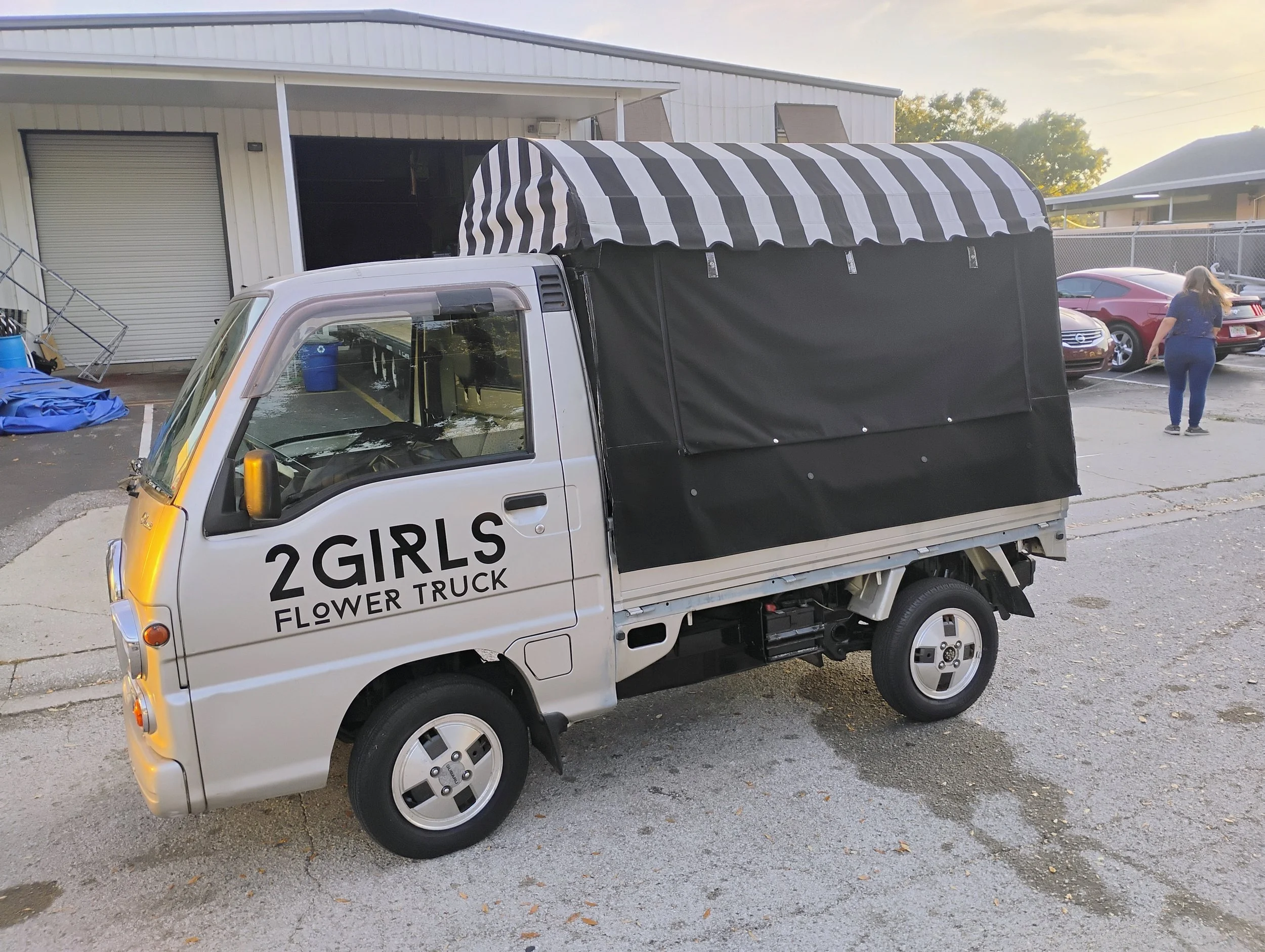 A small silver truck with the words '2 Girls Flower Truck' written on it. The truck has a black and white striped canopy on the back, parked in an outdoor lot near a building, with other cars and a person walking in the background.