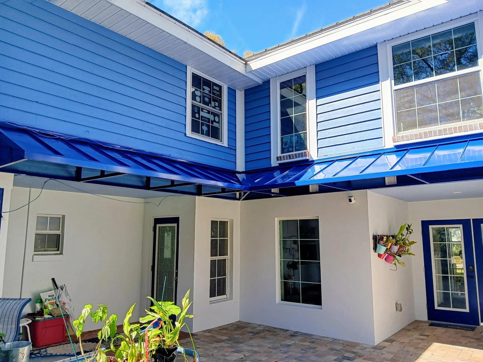 View of a two-story house with blue siding and white trim, featuring multiple windows, a glass door, and a blue metal awning above the patio area. There are plants and outdoor items near the patio.