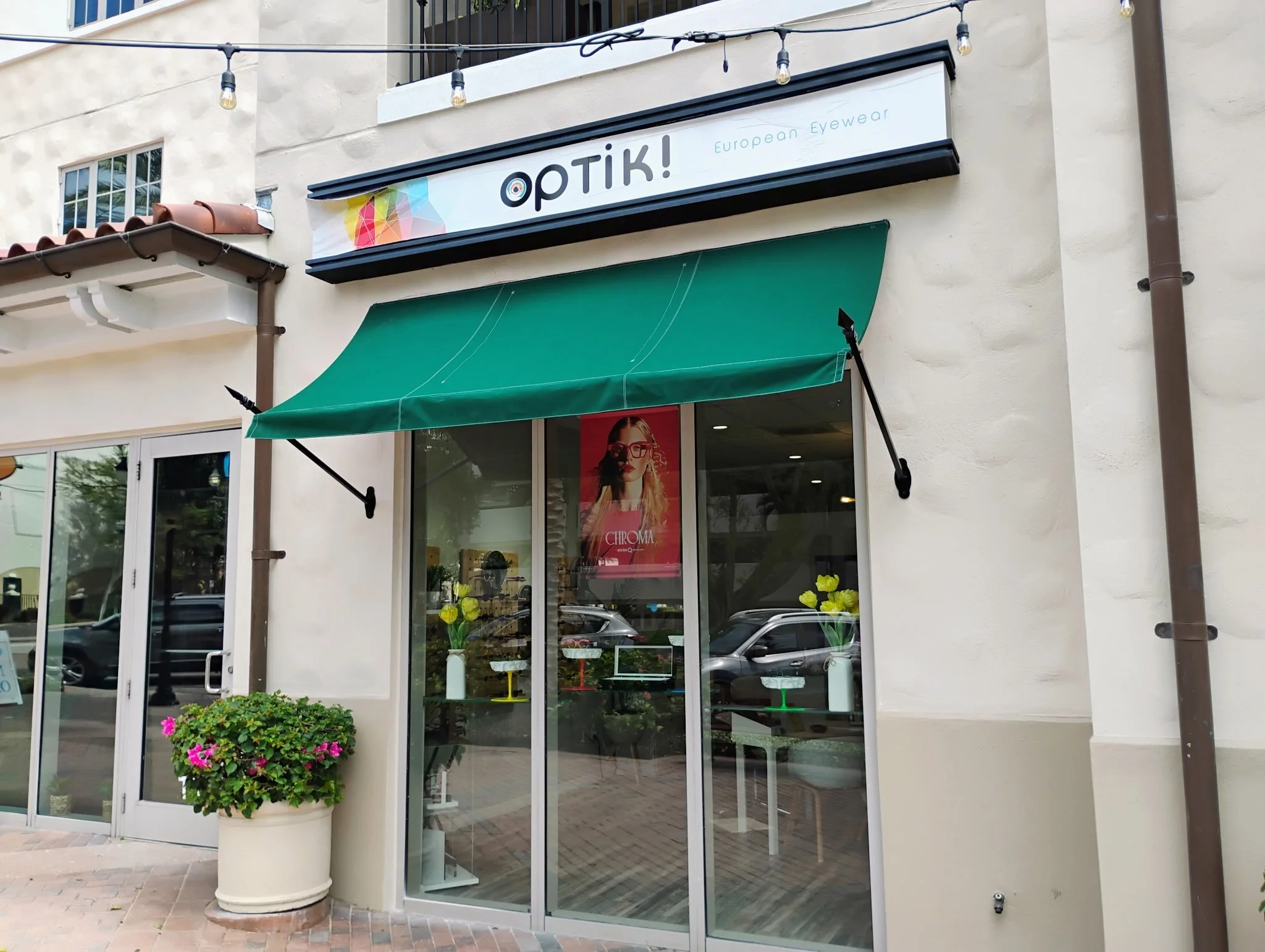 Storefront with a green awning, large glass windows, potted flowers outside, and a sign that reads 'OPTIK! European Eyewear'