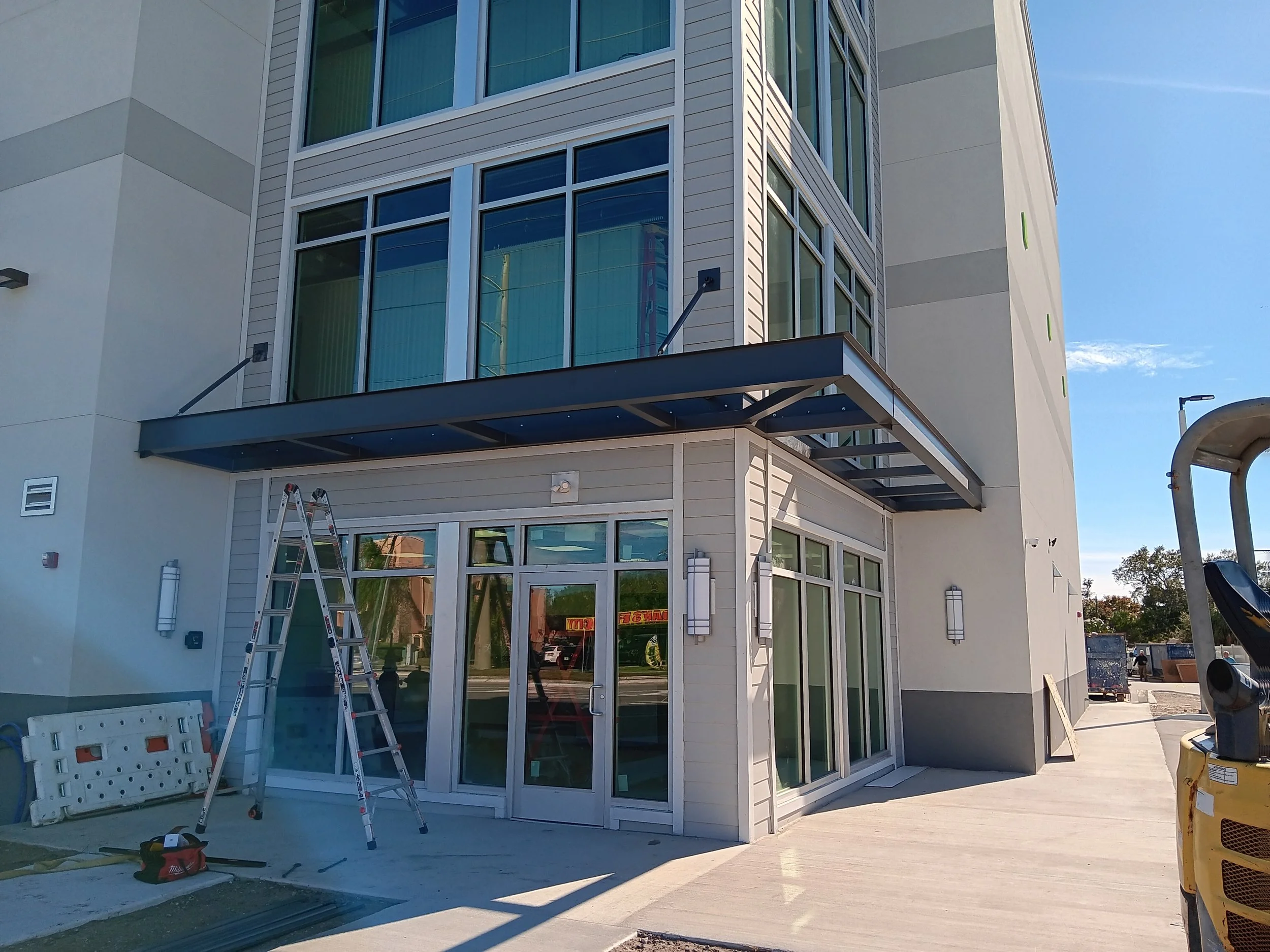 Exterior view of a modern building under construction, featuring large windows, a glass door, a metal awning, and construction tools and scaffolding outside.