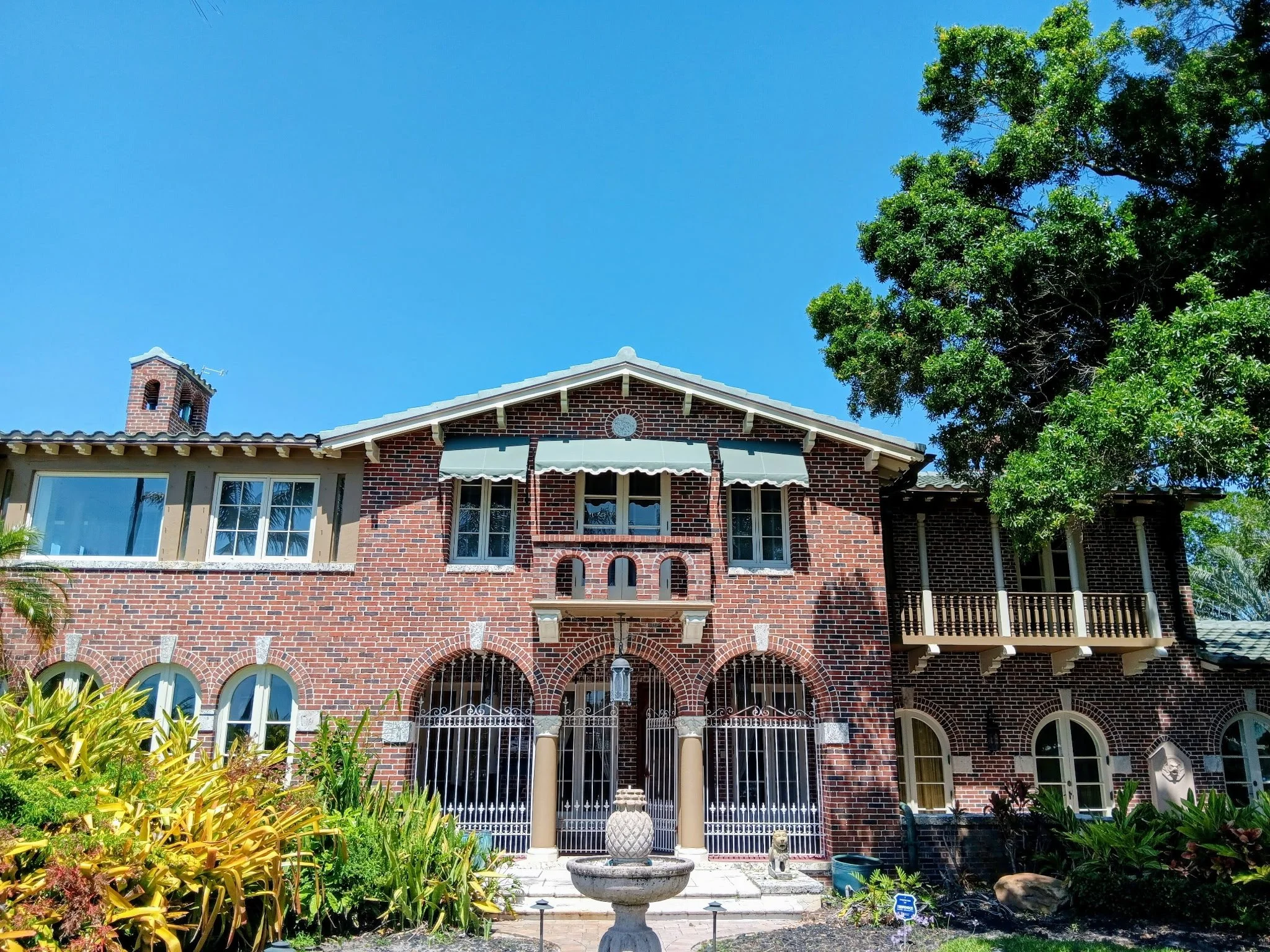 A large brick house with arched windows, a small front garden with plants, and a decorative fountain at the entrance, under a clear blue sky.