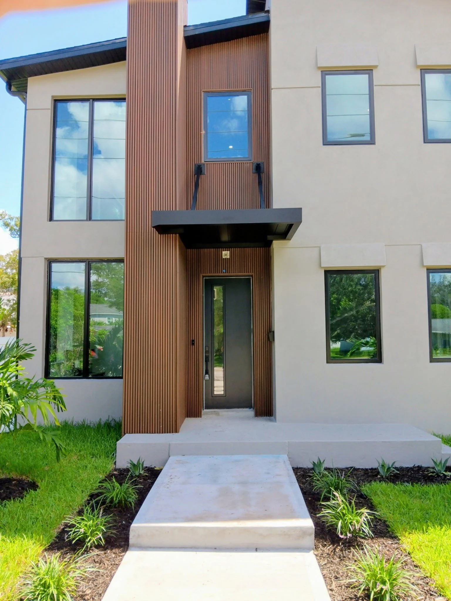 Modern two-story house with a beige exterior, black-framed windows, and a brown vertical wood accent wall above the entrance. Concrete pathway leading to the front door, surrounded by landscaped grass and small plants.