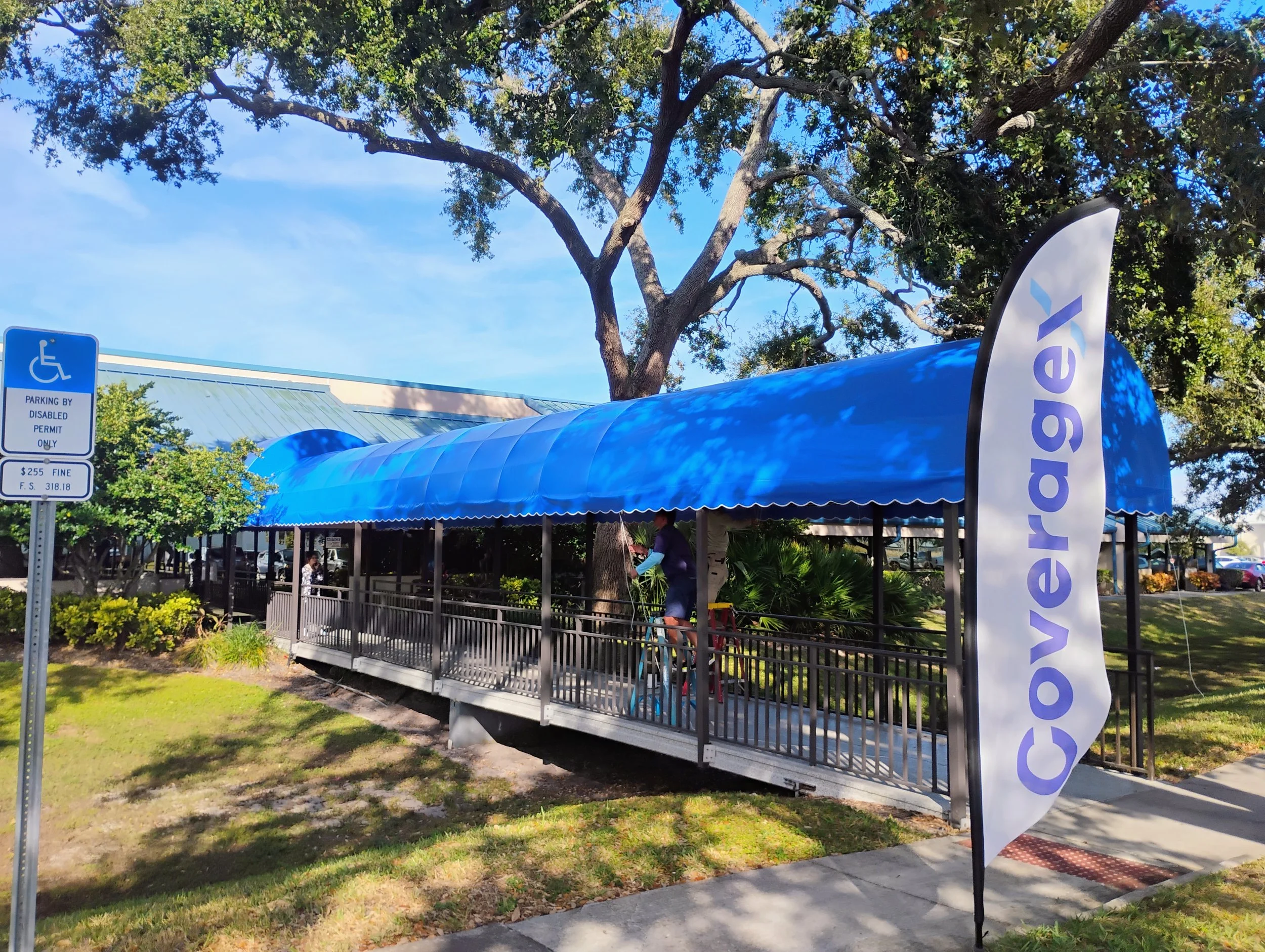 A covered walkway with a blue canopy and a flag with the word 'coveragel' located in a park-like area with trees, plants, and a parking lot in the background. There is also a sign indicating parking for disabled permit only.