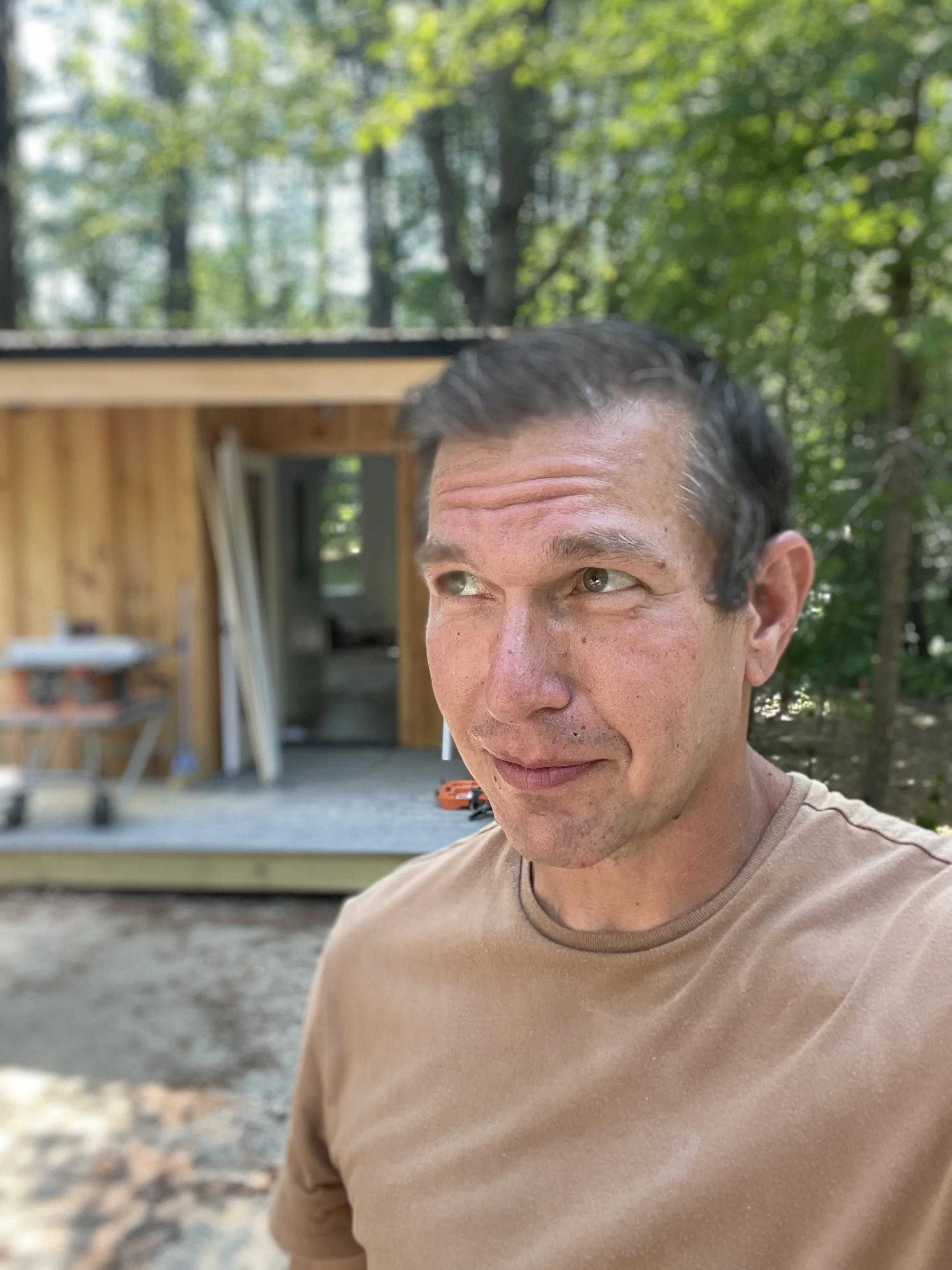 A man with short dark hair and light skin taking a selfie outdoors. Behind him is a partially built wooden structure with trees and greenery in the background.
