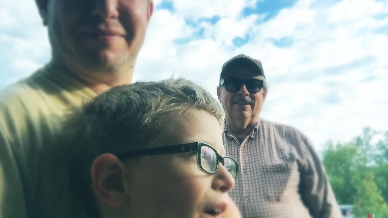 Three people, a young boy in the foreground wearing glasses, an adult man in the background wearing sunglasses and a plaid shirt, and another adult man in the upper left corner partially visible, outdoors on a cloudy day.