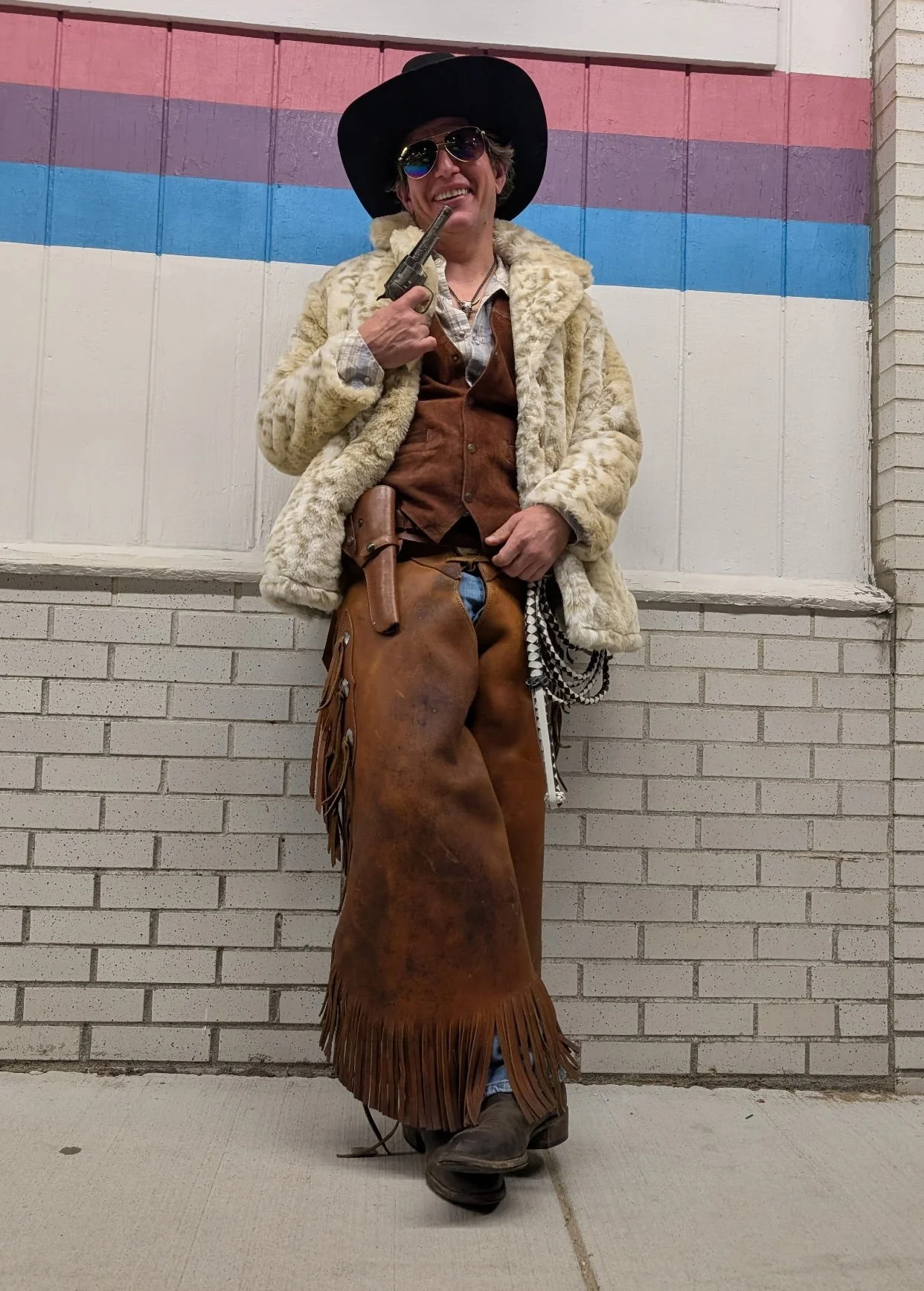 A person dressed in Western-themed costume, wearing a large black hat, aviator sunglasses, a fur coat, cowboy boots, and leather chaps, standing against a wall with a rainbow and transgender pride flag painted on it, and holding a prop revolver near 