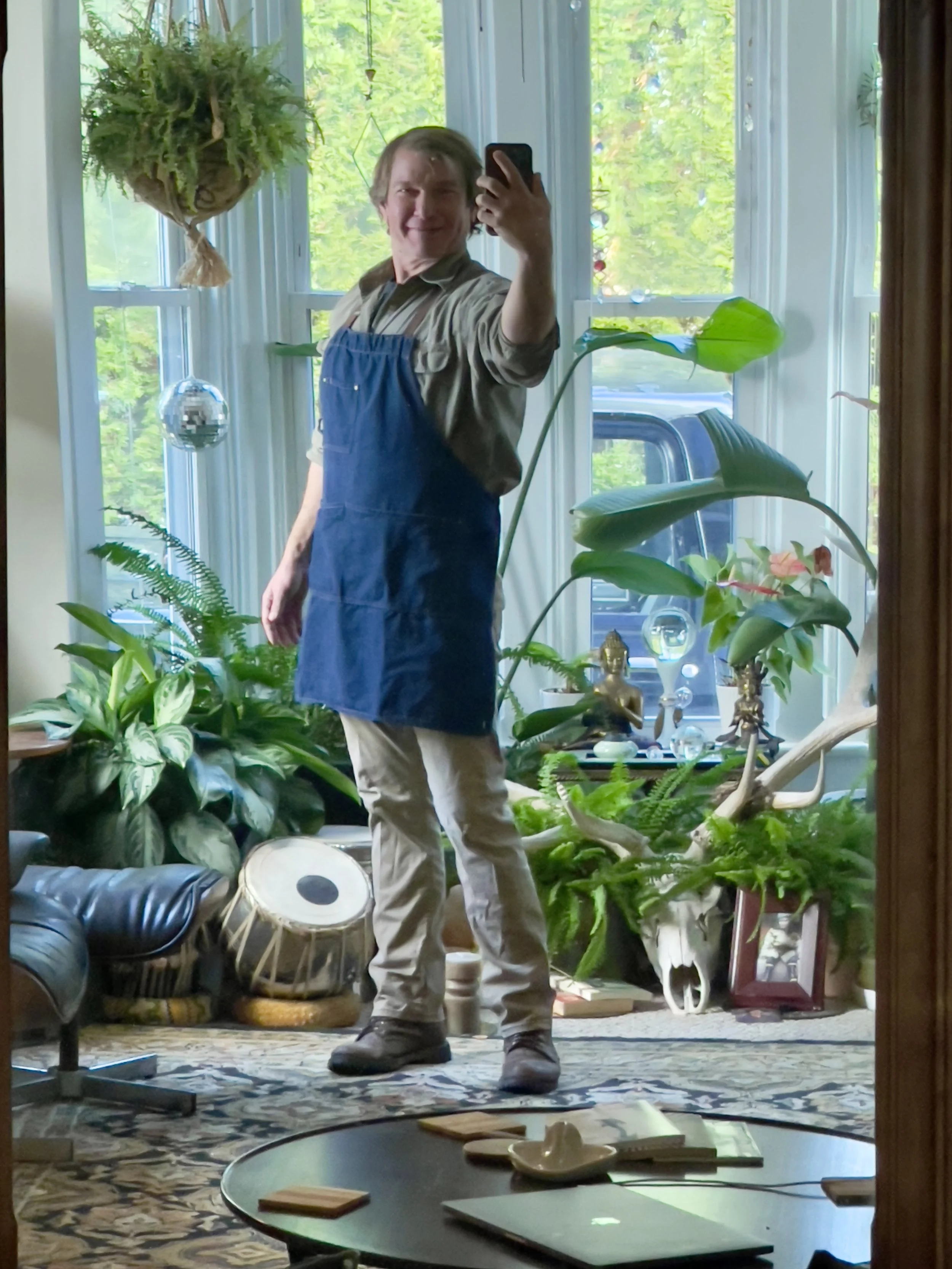 A person taking a selfie in front of a large mirror, surrounded by houseplants and decorative objects in a well-lit room.