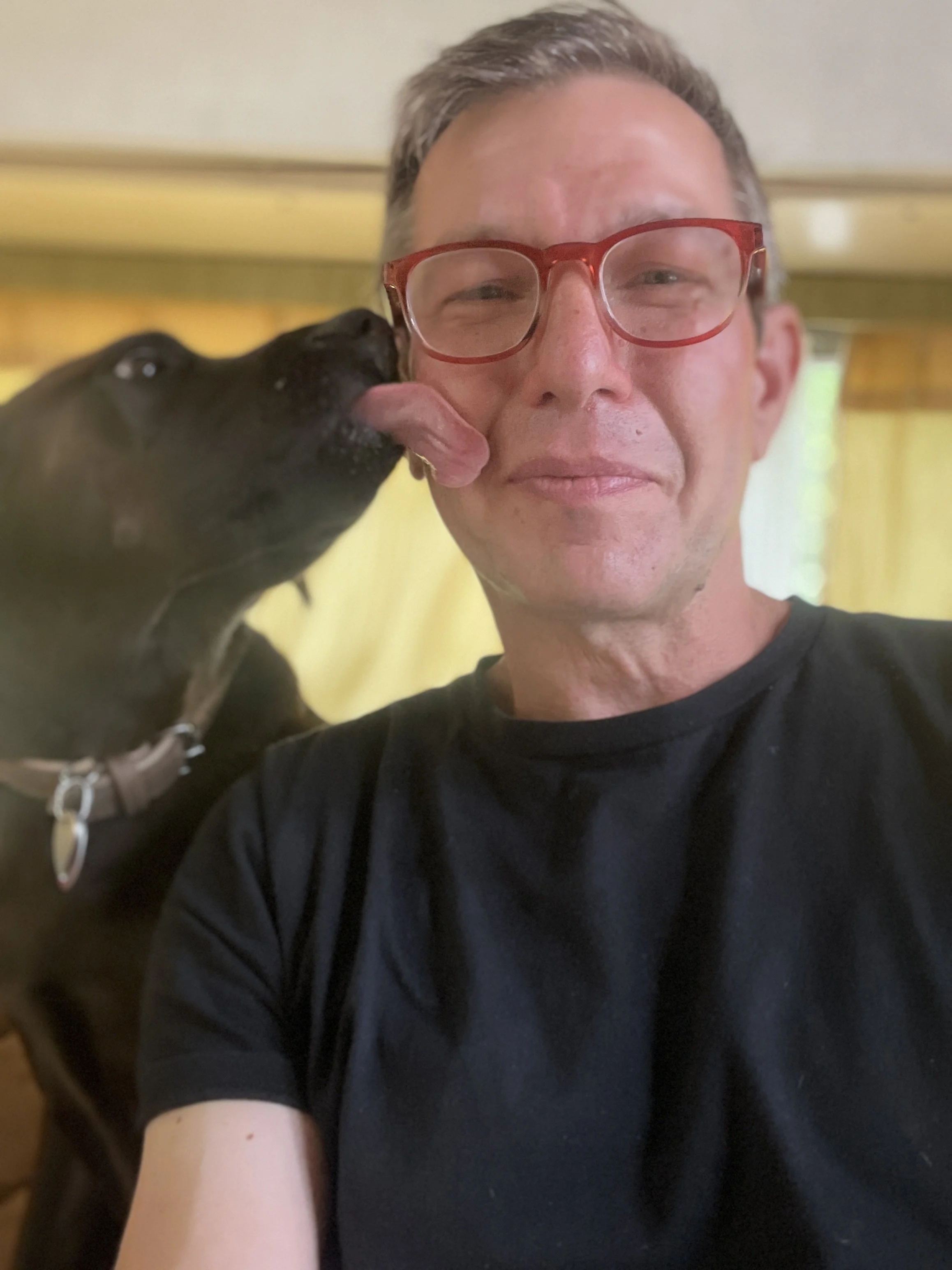 A man with glasses and short gray hair taking a selfie with a black dog licking his cheek. The background has yellow curtains.