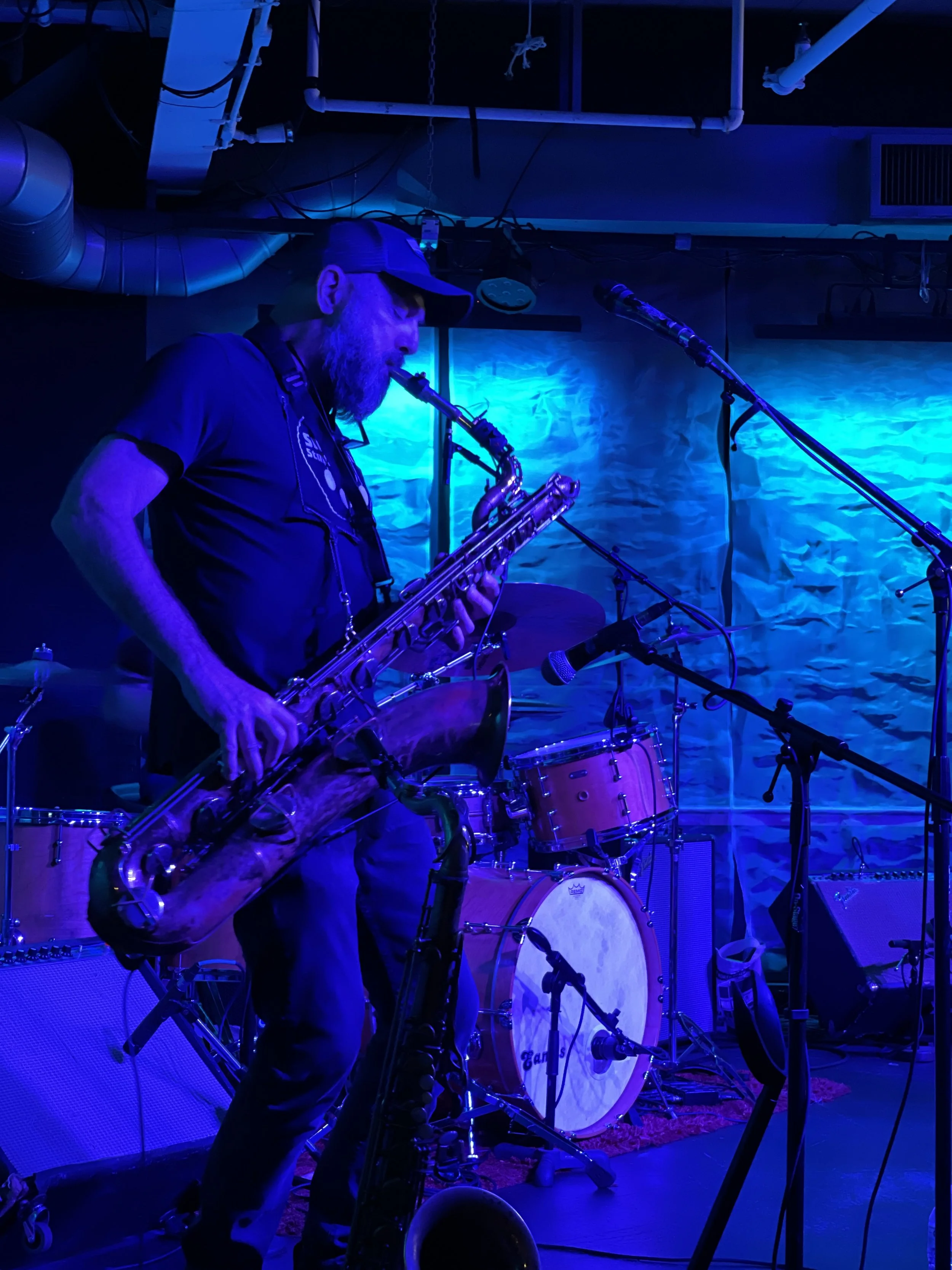 A musician playing a tenor saxophone on stage, illuminated by blue stage lighting, with a drum set and microphones visible in the background.