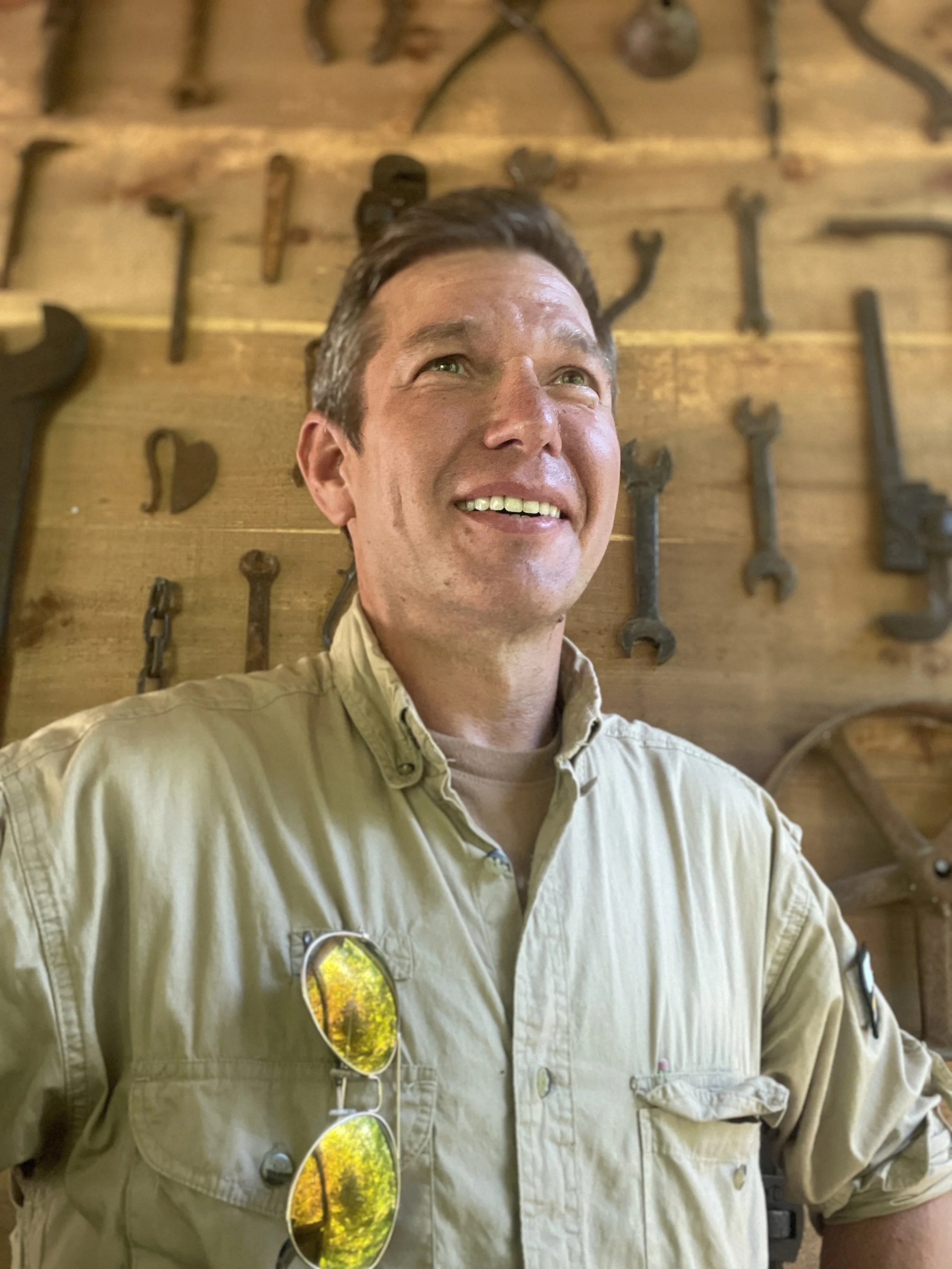 A man with brown hair smiling, wearing a beige shirt with sunglasses hanging from the front, standing in front of a wooden wall decorated with vintage tools.