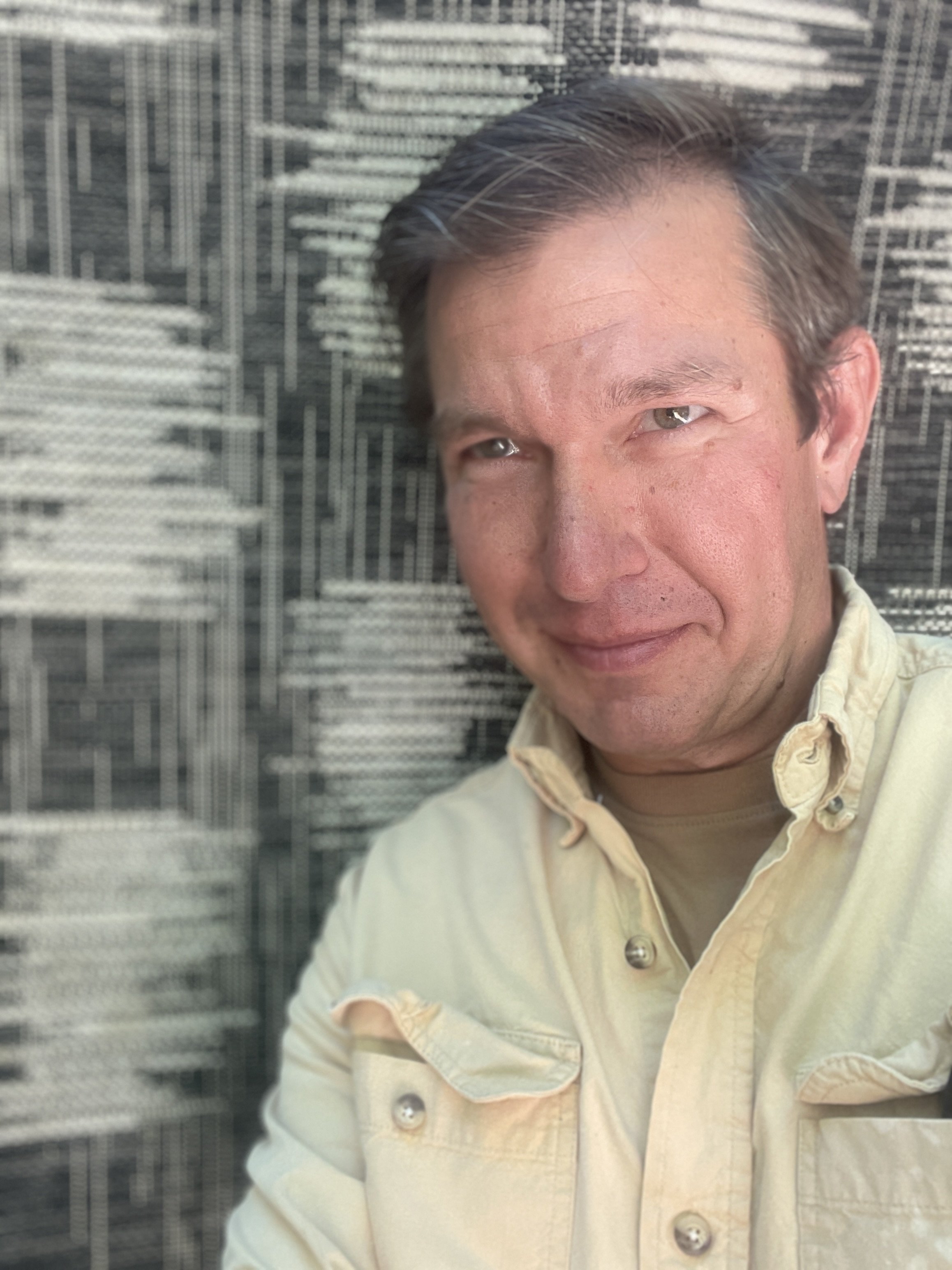 A man with short brown hair and light skin, smiling and wearing a light beige shirt, is taking a selfie against a background of a black and white cityscape artwork.