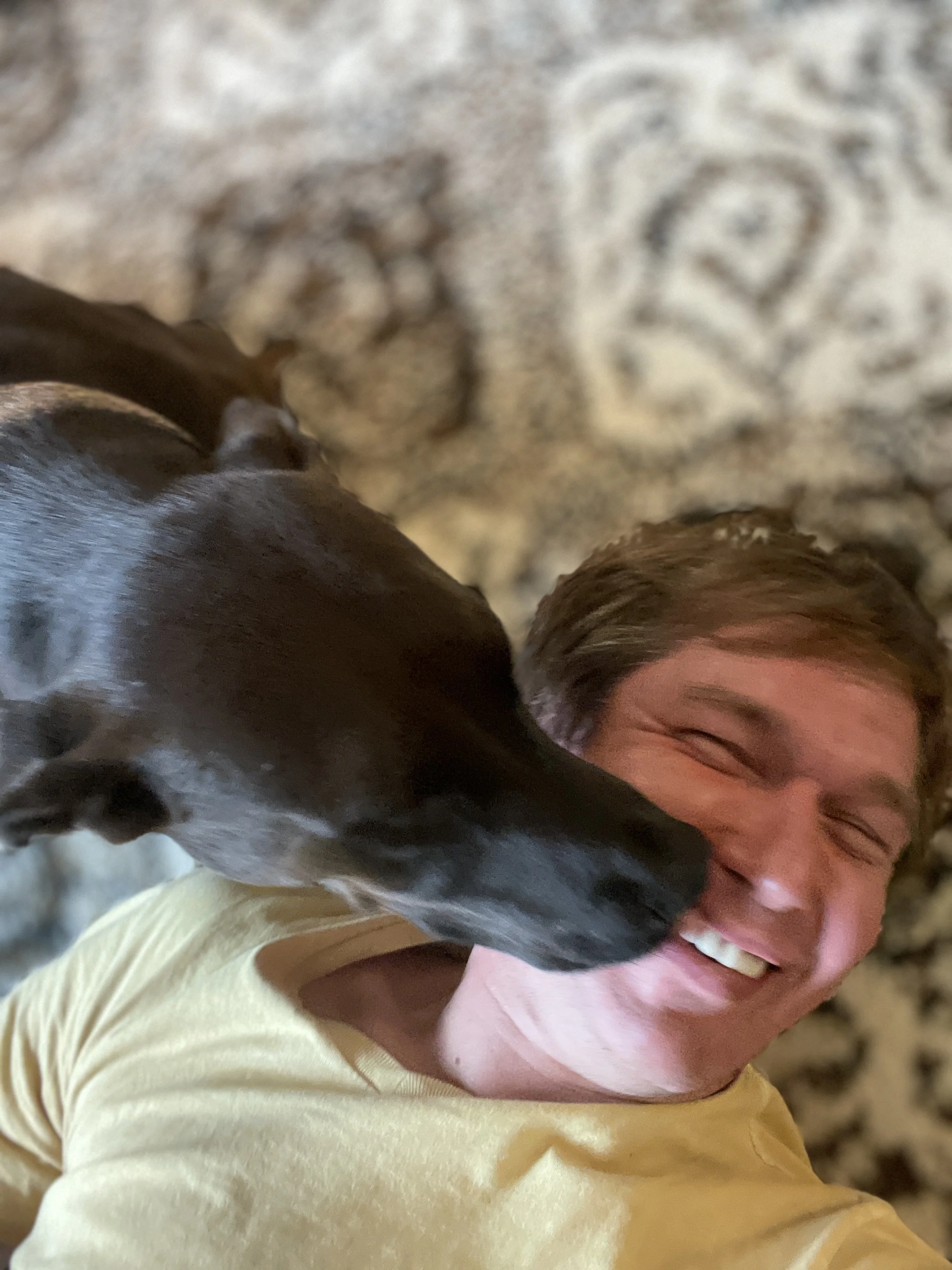 A person with short hair smiling with eyes closed, wearing a yellow shirt, being licked on the face by a black and white dog.