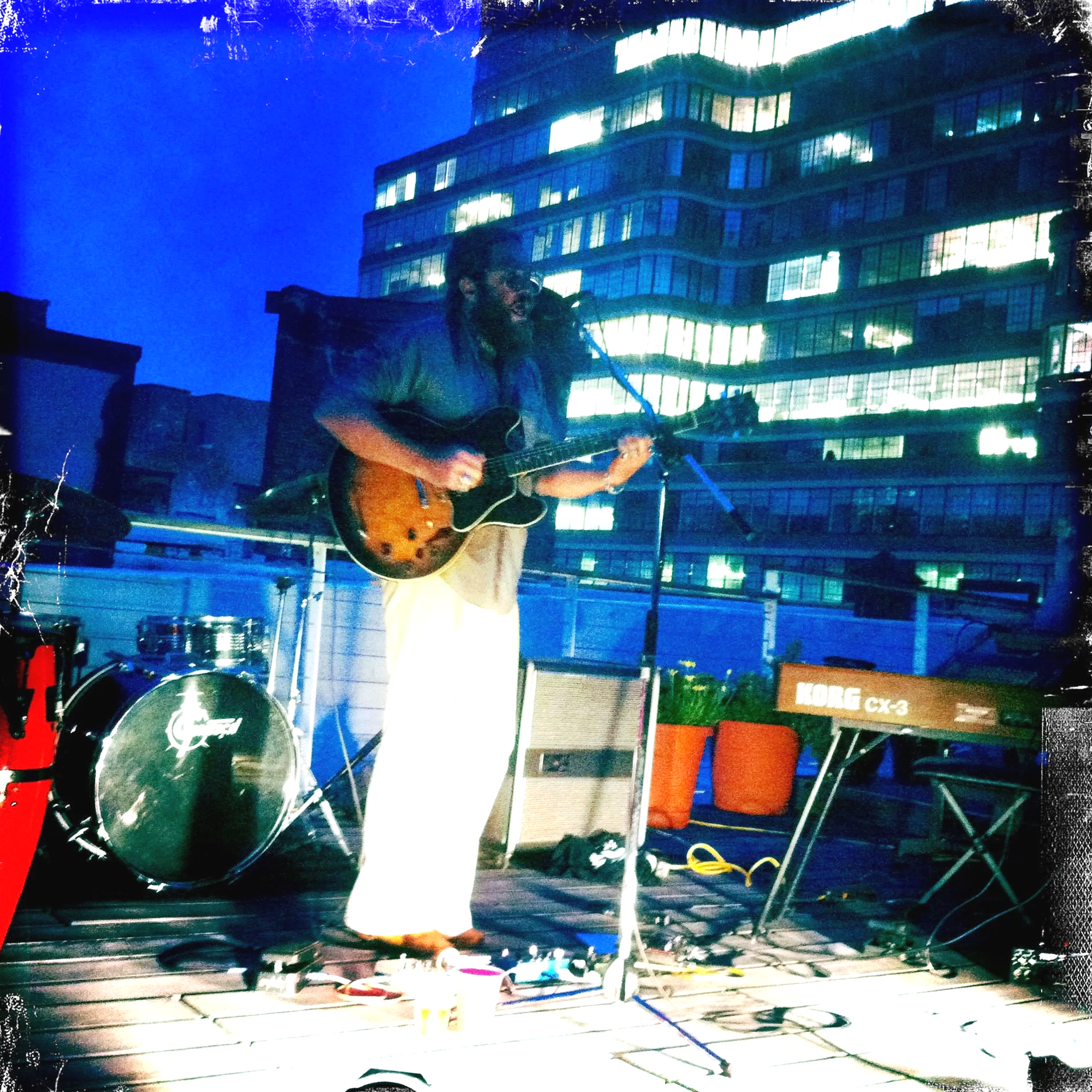 Musician Bing Ji Ling playing an electric guitar and singing at private party at thelab, with  NYC cityscape of illuminated office buildings in the background. There is a drum set and a keyboard nearby. Photo By Julian Schlaver