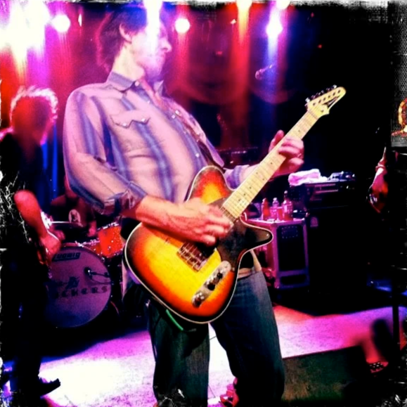 Cooley from the Drive By Truckers playing an electric guitar on stage with colorful lighting, with drummer and other band members in the background. Photo By Julian Schlaver