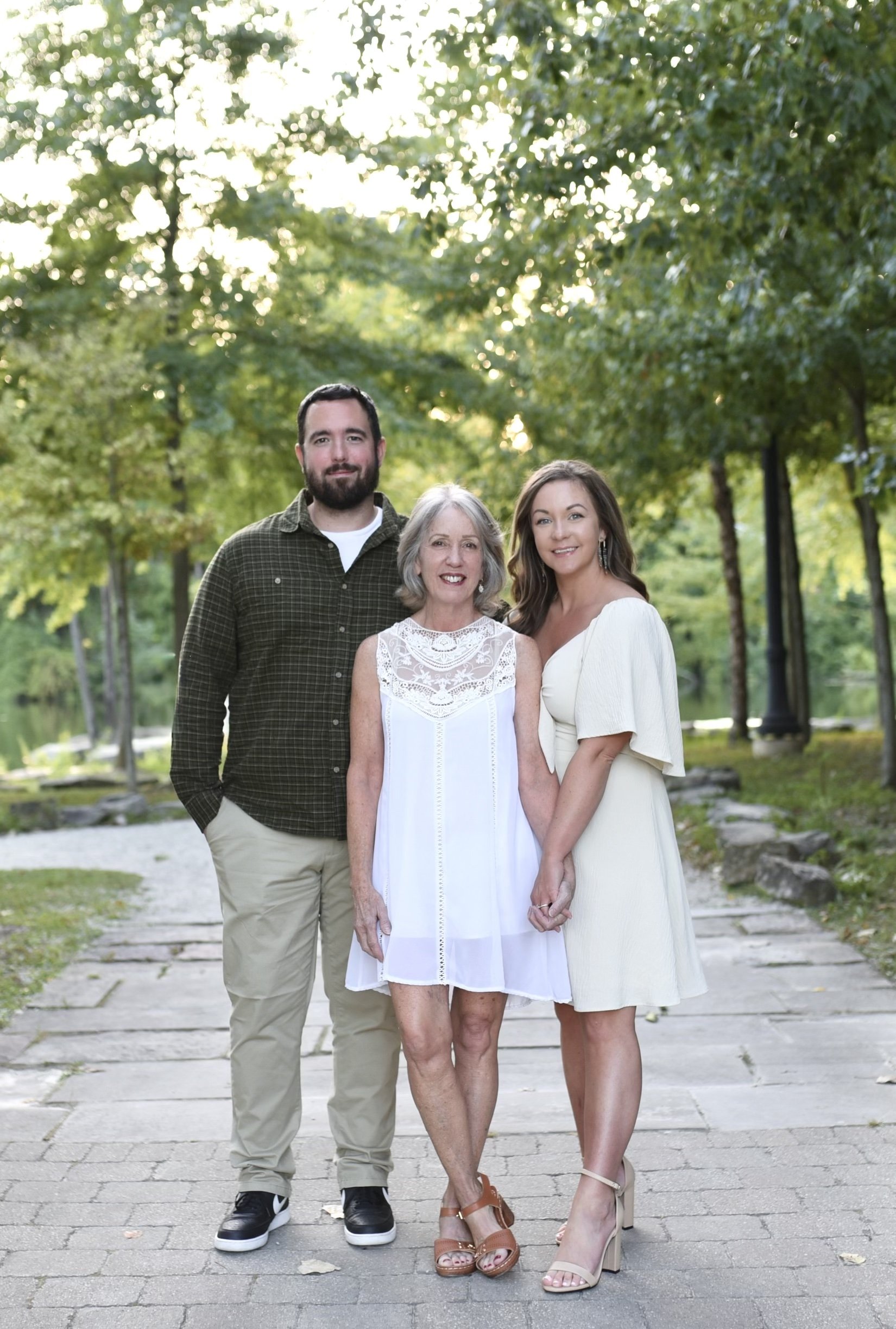 A family of three standing on a stone path in a park with trees, smiling at the camera.