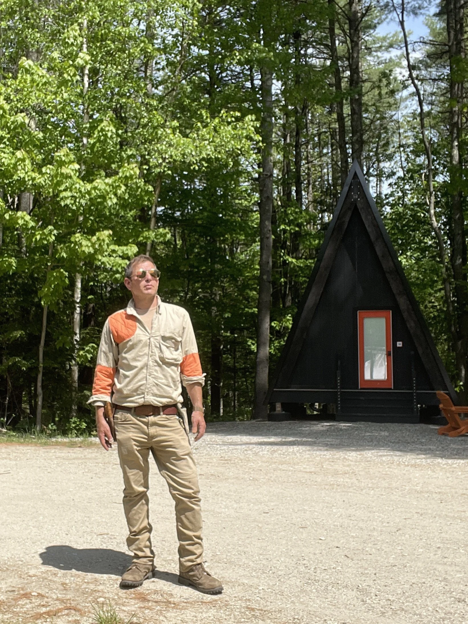 A man wearing khaki pants, a beige jacket with orange patches, and sunglasses standing outdoors in a wooded area near a small black triangular building with a red door.