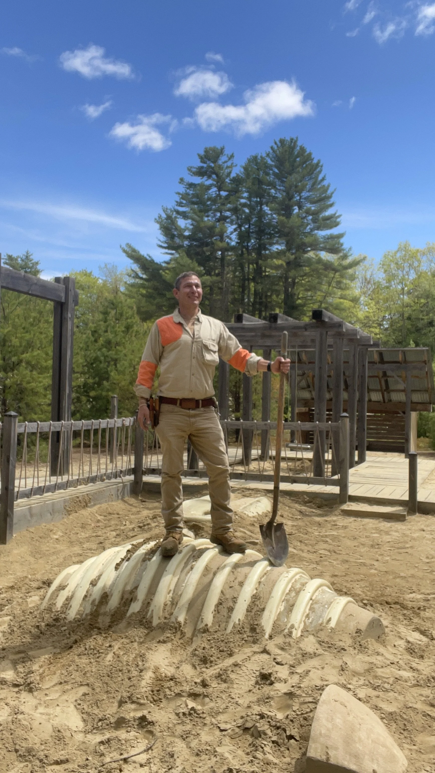 A person standing on a large dinosaur fossil, holding a shovel, outdoors on a sunny day with trees and a wooden structure in the background.
