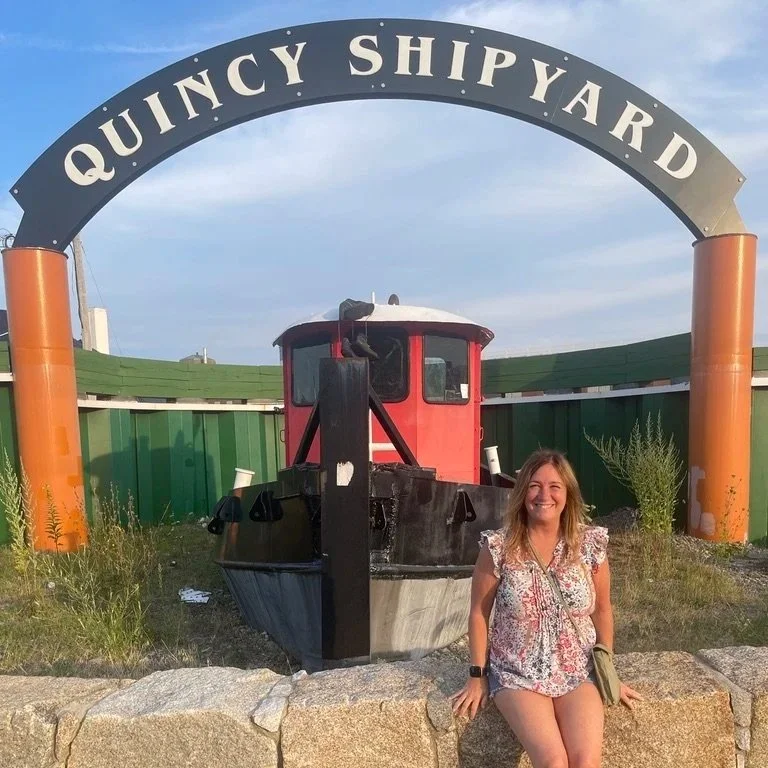 A smiling woman sitting on a stone wall in front of a boat display at Quincy Shipyard, with a large sign overhead that reads 'QUINCY SHIPYARD'.