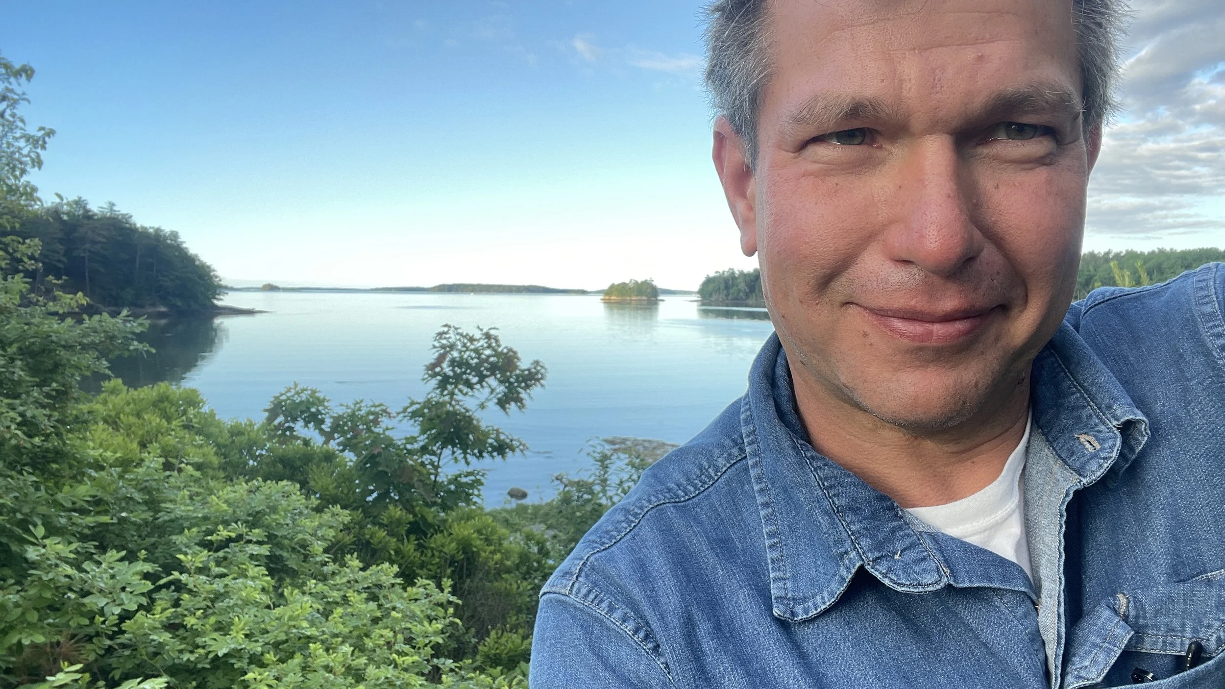 A man smiling in front of a lake with trees and small islands, wearing a denim jacket and a white shirt, on a clear day with some clouds.