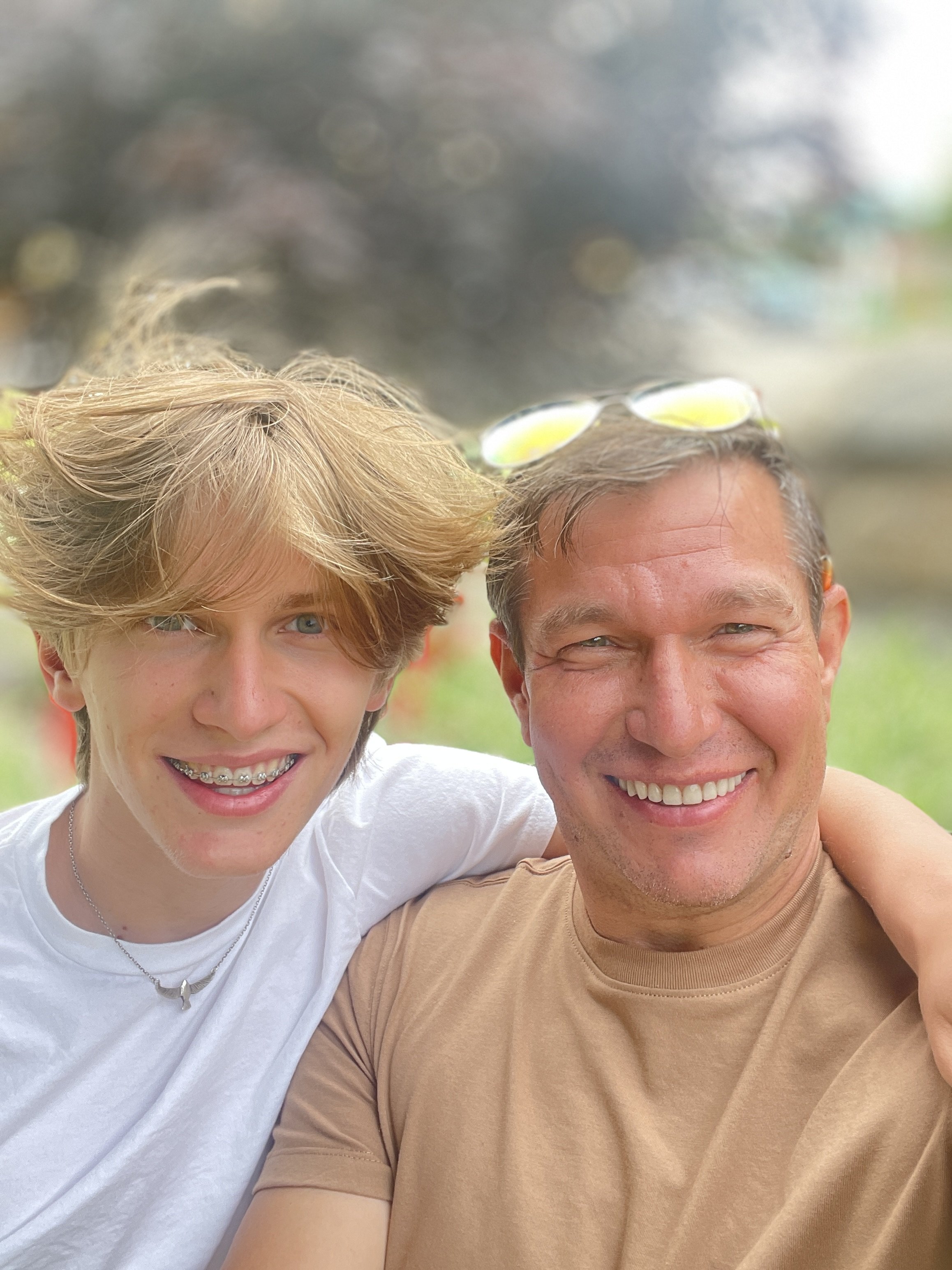 A smiling, named Julian Schlaver, man and a smiling young person with braces taking a selfie outdoors, with a blurred background and sunglasses on the man's head.