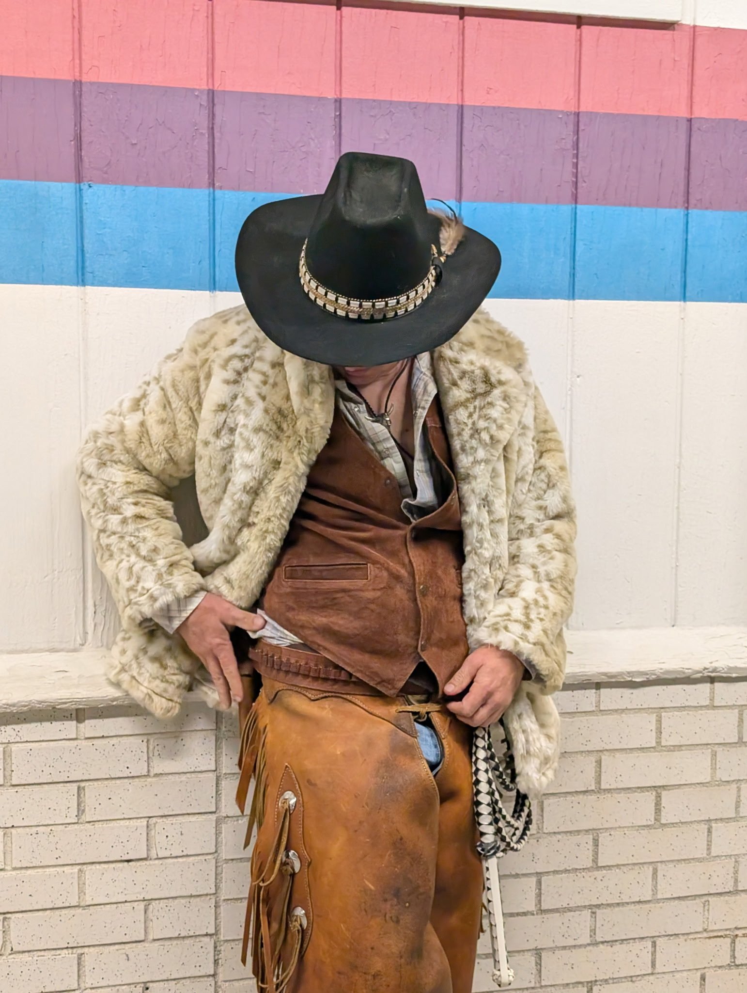 Person wearing a black cowboy hat, a beige faux fur coat, brown leather chaps, and a plaid shirt, leaning against a brick wall with pastel-colored stripes.