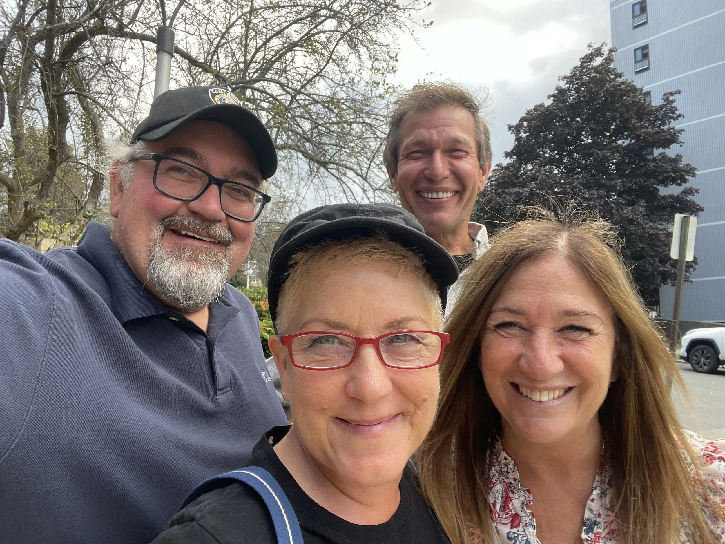 Group of five people smiling outdoors, trees, cloudy sky, and buildings in background.