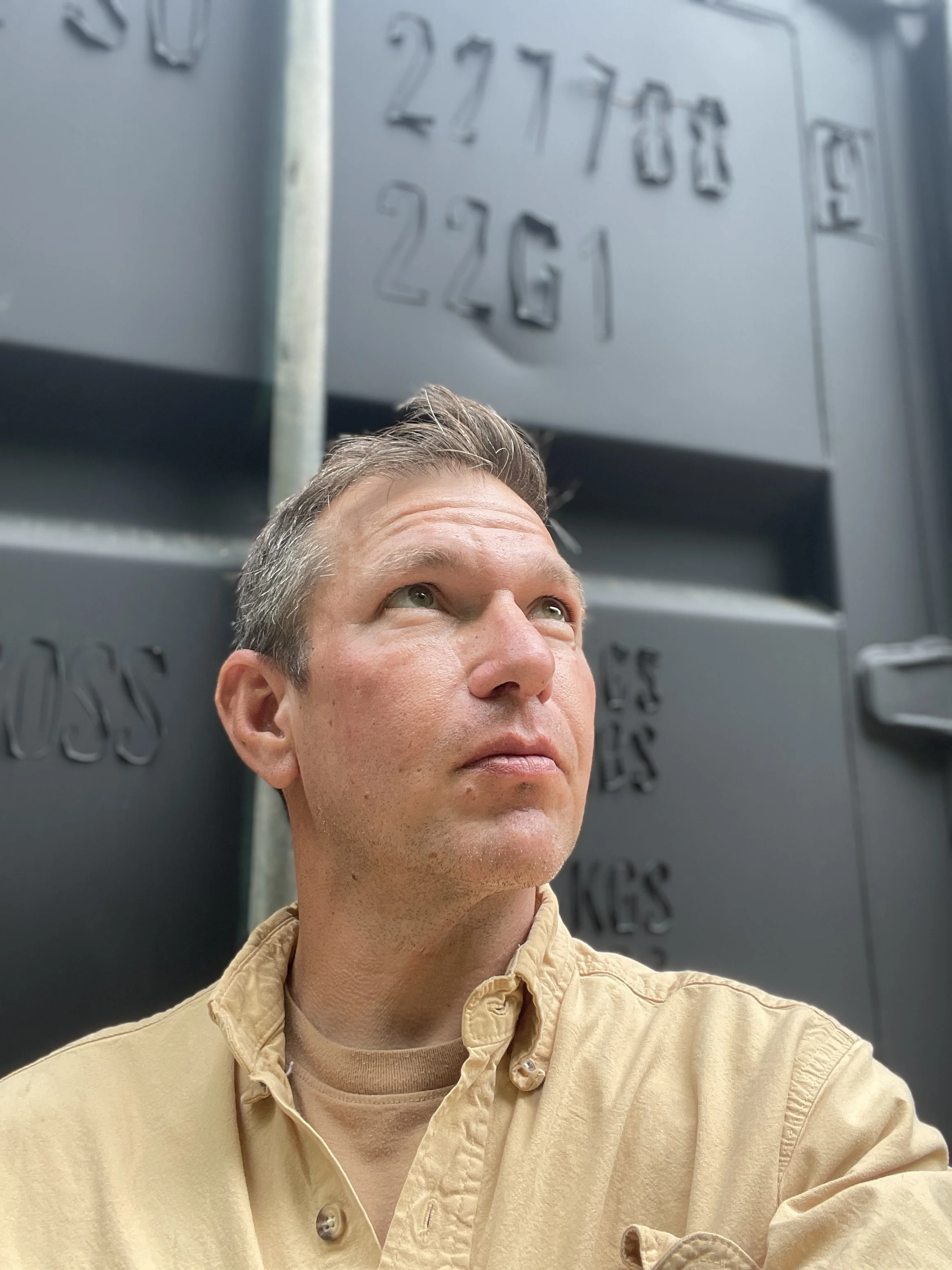 A man with short, styled hair looking upward against a background of dark gray electrical boxes or panels with embossed numbers and letters.