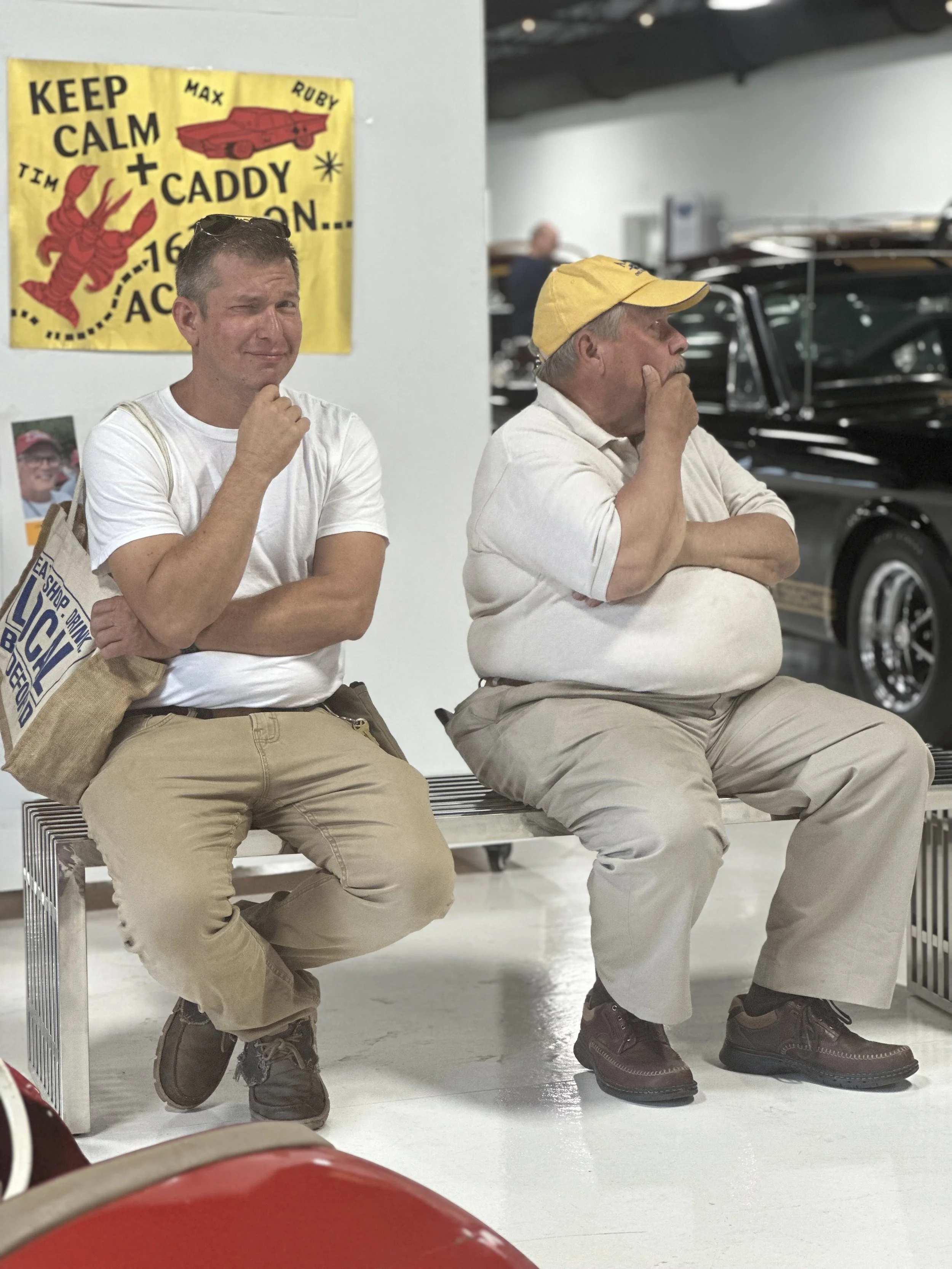 Two men sit on a bench, one looking at the camera with a slight smile and the other in profile with his hand on his chin. There is a yellow poster on the wall behind them that says "KEEP CALM" with a lobster, a car, and other decorative elements. The